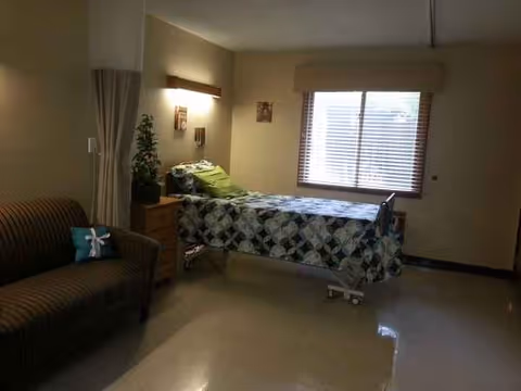 Senior living facility bedroom featuring a single bed, quilt, nightstand, sofa, and window.