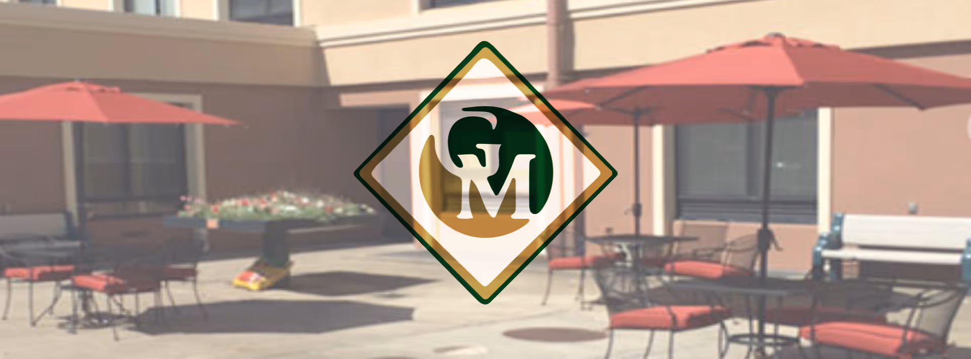 Outdoor courtyard with patio tables and red umbrellas in front of a building, overlaid by a translucent 'GM' logo.