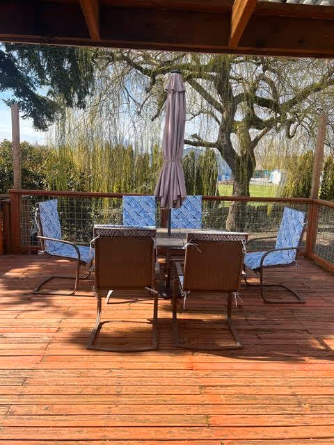 Outdoor wooden deck with a glass-top table surrounded by six chairs, four with blue cushions and two without. A closed beige patio umbrella is centered on the table. In the background, there are large trees and greenery under a clear sky.