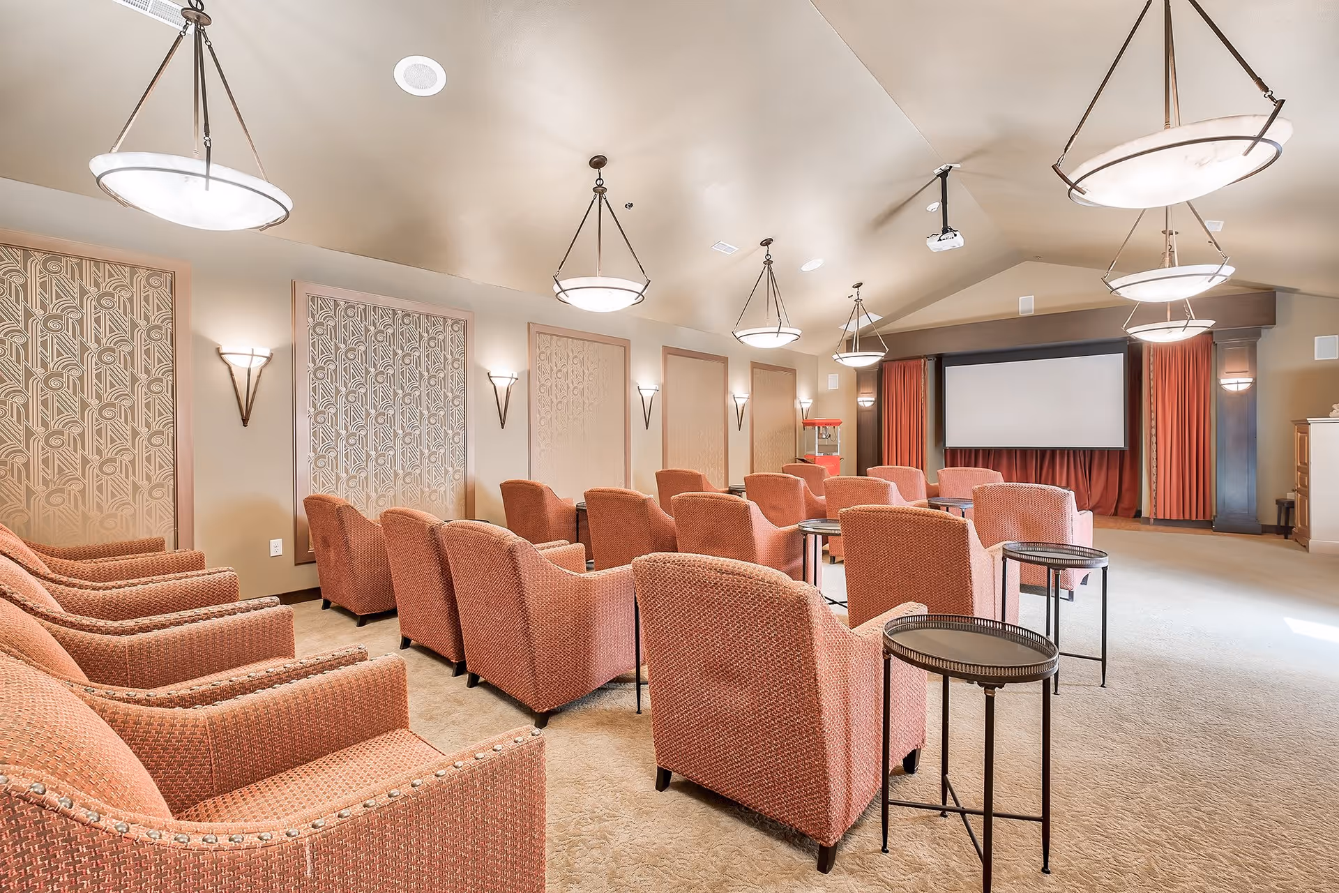 A cozy theater room with multiple rows of cushioned armchairs facing a large projection screen. The room features warm lighting from ceiling fixtures and wall sconces, decorative wall panels, and a popcorn machine near the screen.