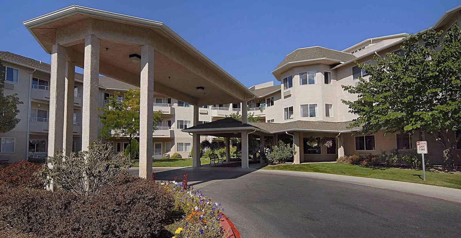 Covered entrance driveway and porte-cochère of a multi-story beige senior living building with landscaping and flower beds.