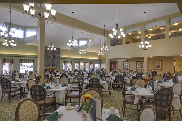 A spacious dining room with multiple round tables covered with white tablecloths, each set with green napkins, cups, and silverware. The room features high ceilings with several chandeliers and large windows letting in natural light. There is a stone fireplace on one side and framed artwork on the walls.
