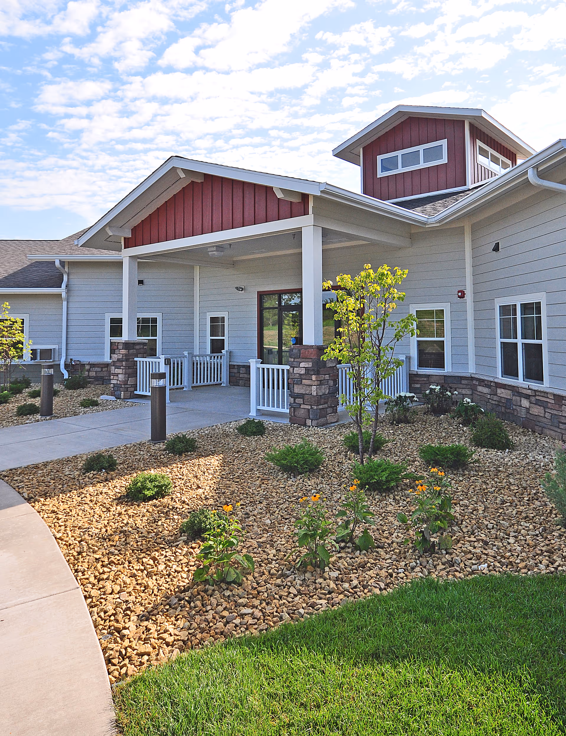 Exterior view of the entrance to Spring Valley Senior Living and Health Care Campus, featuring a covered porch with white railings, stone pillars, and a landscaped garden with small bushes and flowers under a partly cloudy sky.