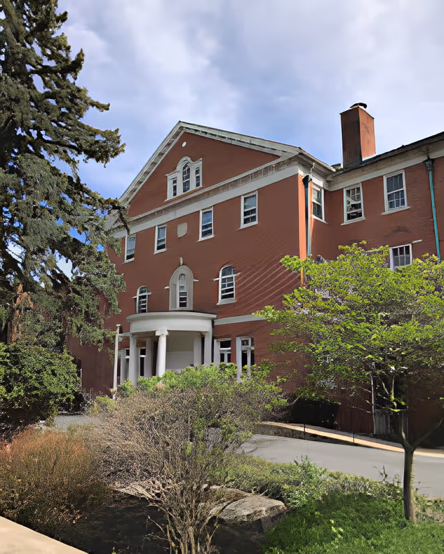 Exterior view of a large, red brick senior living facility building with white trim and columns at the entrance. The building has multiple windows and a chimney. Surrounding the building are trees, bushes, and landscaped greenery under a partly cloudy sky.