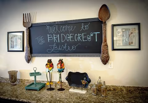 A kitchen or dining area countertop with decorative items including two colorful bird figurines, a small blackboard sign, and various bottles. Above the countertop is a chalkboard framed by a large wooden fork and spoon, with the handwritten message 'Welcome to the Bridgecrest Bistro'. Two framed pictures hang on the wall on either side of the chalkboard.
