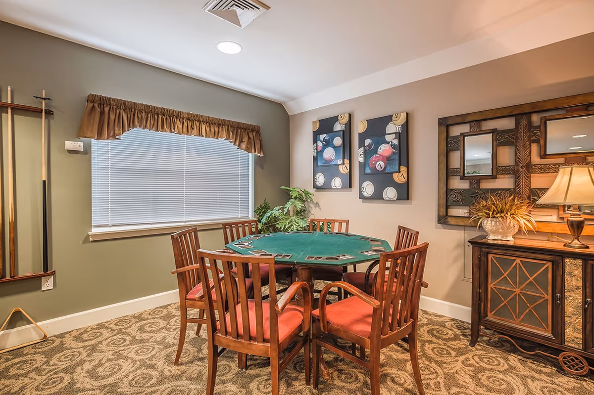 A cozy game room with a round green felt card table surrounded by six wooden chairs with red cushions. The room has patterned carpet, a window with blinds and a brown valance, two paintings of billiard balls on the wall, a decorative mirror, a lamp on a wooden cabinet, and a rack holding pool cues in the corner.