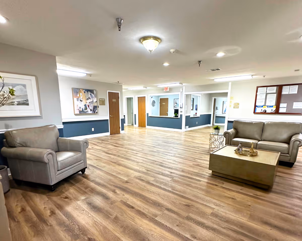A spacious and well-lit common area with wooden flooring, featuring a gray armchair on the left and a gray sofa on the right. A wooden coffee table with decorative items is placed in front of the sofa. The walls are painted white with a blue accent panel, and there are framed artworks and a bulletin board on the walls. Several doors and a reception window are visible in the background.