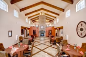 Spacious assisted living dining room with multiple round tables covered with brown tablecloths and pink napkins, cushioned chairs, a high vaulted ceiling with wooden beams, large windows near the ceiling, and a stone fireplace at the far end of the room.