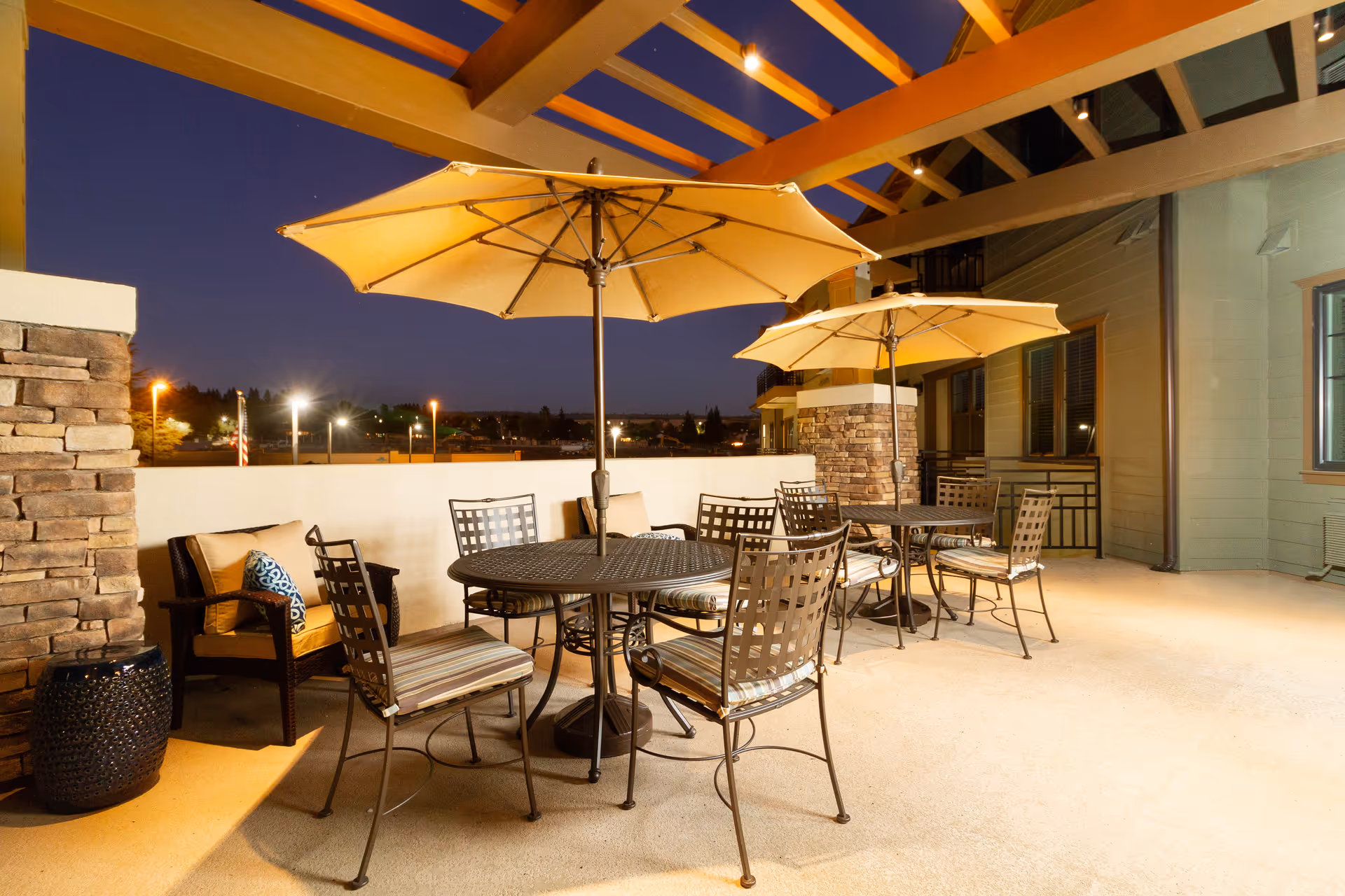 Outdoor patio area at night with two round metal tables, each with four chairs and a large beige umbrella. There is a cushioned armchair with pillows against a stone pillar, and the patio is covered by a wooden pergola. Lights and buildings are visible in the background.