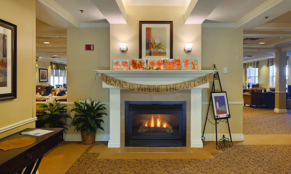 Lit fireplace in a senior living common room with artwork on the mantel, a banner, potted plants, and seating visible in the background.