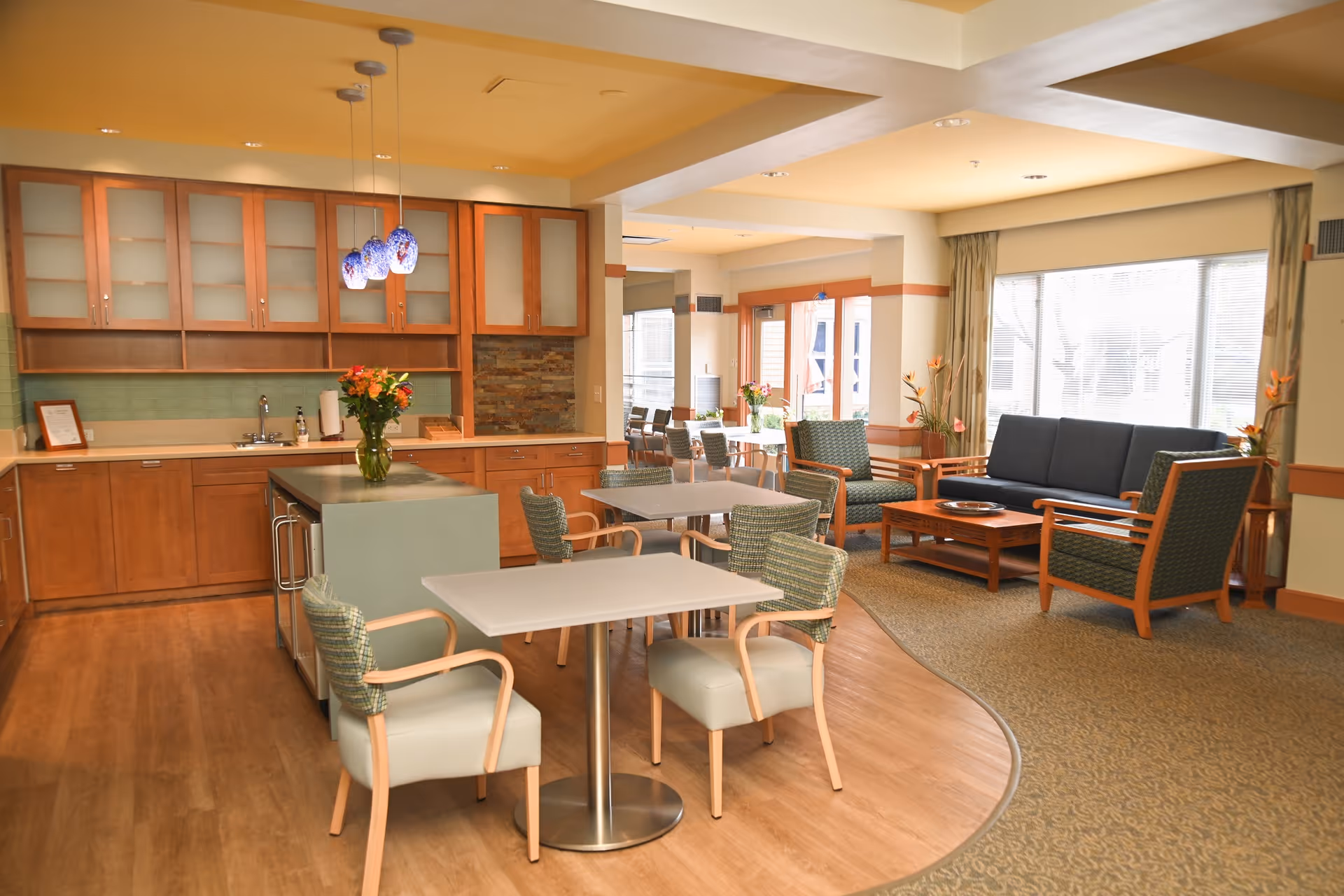 A bright and spacious common area in a senior living facility featuring a kitchen with wooden cabinets, a small island with a vase of flowers, and a dining area with tables and chairs. Adjacent to the dining area is a living room space with a sofa, armchairs, a coffee table, and large windows letting in natural light.