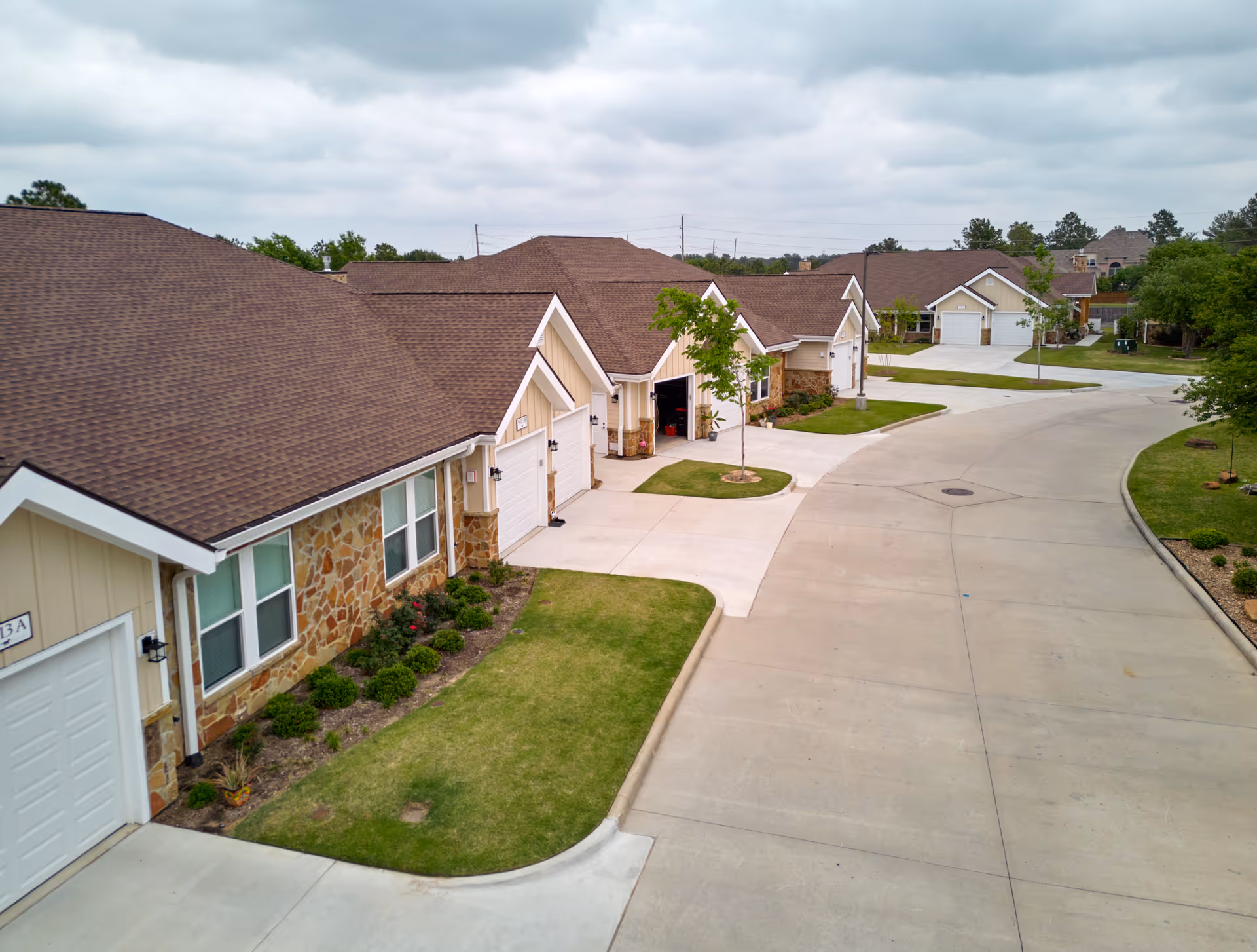 A row of single-story residential buildings with beige siding and stone accents, each with a garage and small front lawn, situated along a curved concrete driveway under a cloudy sky.