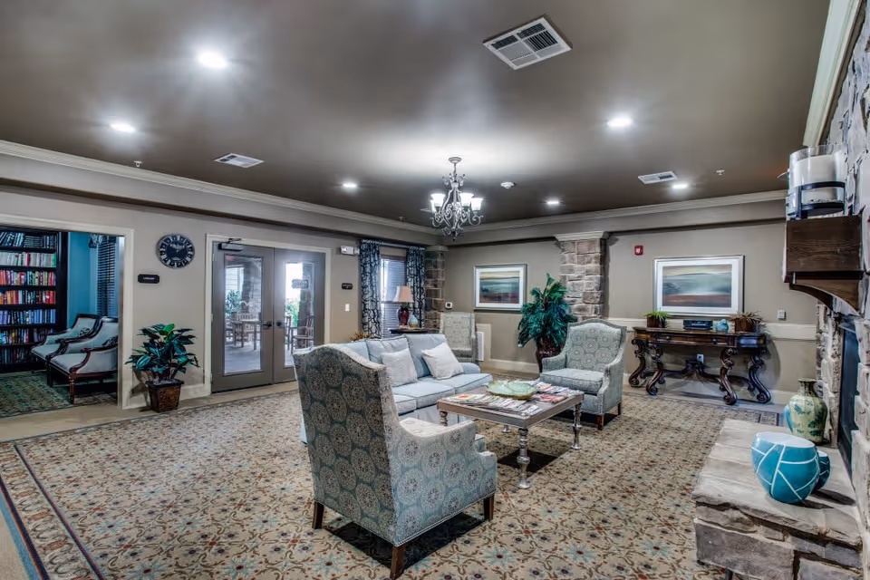 A cozy living room area in a senior living facility featuring a patterned carpet, a sofa with cushions, two upholstered armchairs, a wooden coffee table with magazines, and a stone fireplace with decorative vases. The room has beige walls with stone accents, framed artwork, a chandelier, and large glass doors leading to an outdoor patio. There is also a side table with a lamp and a potted plant near the entrance to a library room filled with bookshelves and chairs.