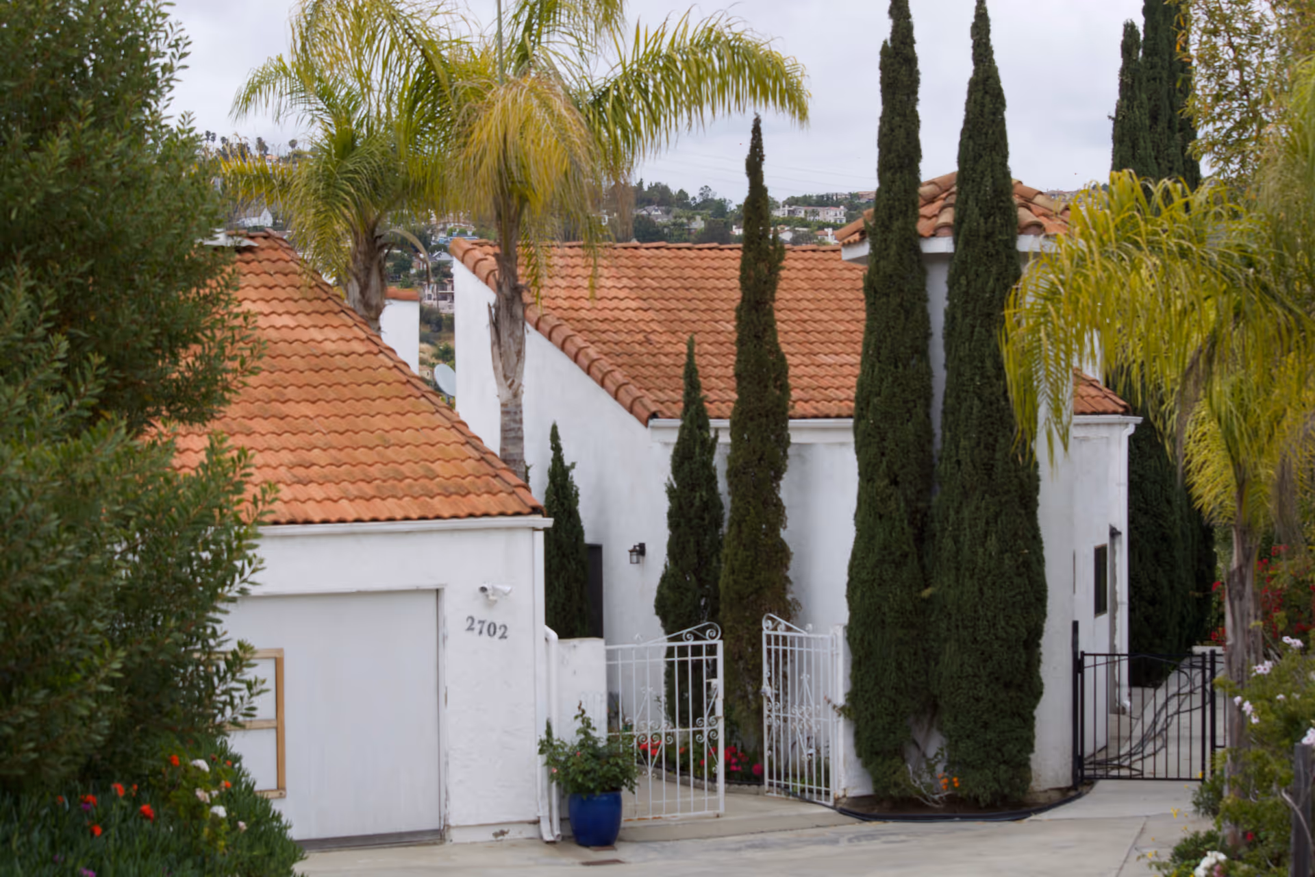 Exterior view of a white building with red tile roofs surrounded by tall cypress trees and palm trees. There is a white metal gate and a driveway leading to a garage door with the number 2702 on it. The sky is overcast.
