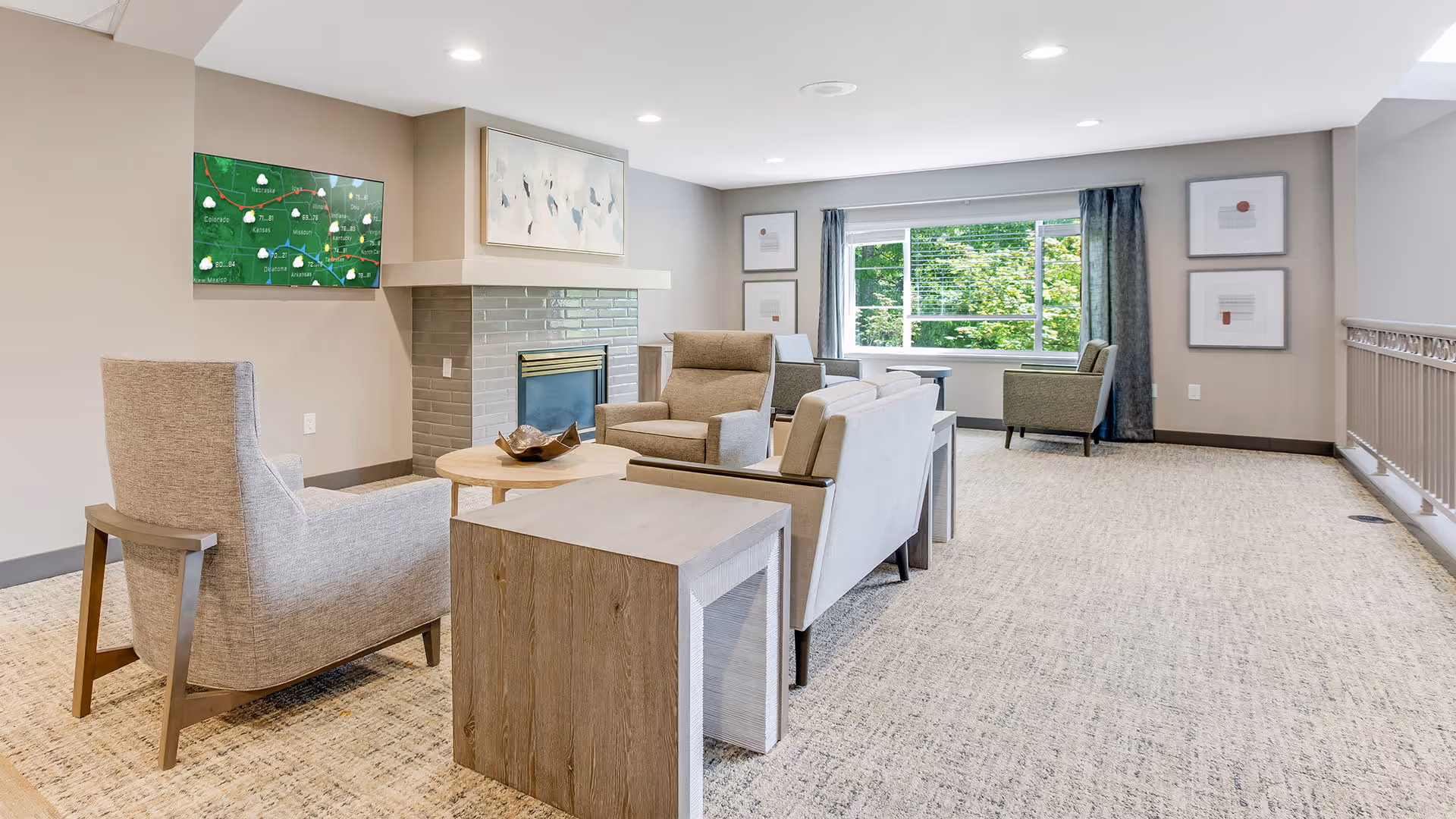 A bright and spacious living room area with beige walls and carpeted floor. The room features a fireplace with a painting above it, a wall-mounted TV displaying a weather map, and several comfortable armchairs and sofas arranged around a wooden coffee table. Large window with curtains lets in natural light and offers a view of greenery outside. Framed artwork is hung on the walls.