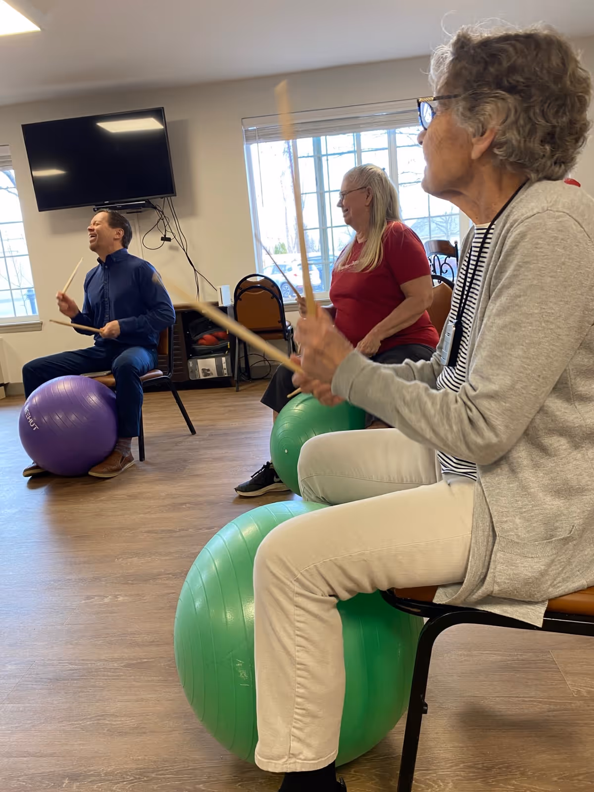 Three elderly people sitting on chairs with large exercise balls between their legs, holding drumsticks and participating in a seated drumming exercise in a bright room with large windows and a wall-mounted TV.