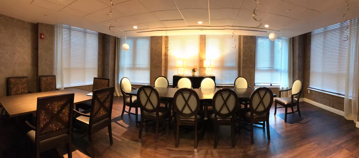 A well-lit dining room with two large tables surrounded by multiple chairs. The room has large windows with blinds, two table lamps on a sideboard, and decorative hanging streamers from the ceiling. The floor is wooden, and the walls have a patterned wallpaper.