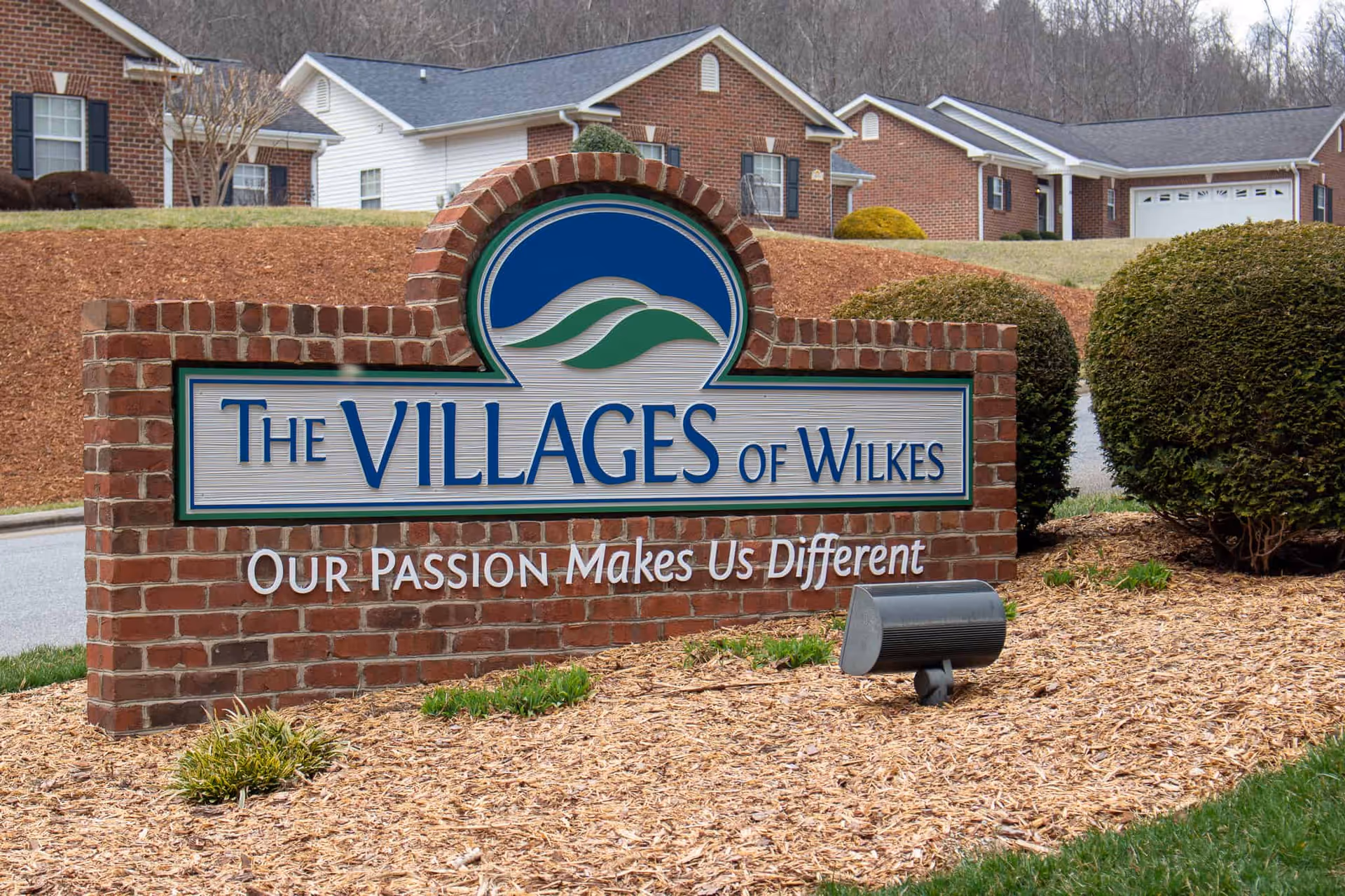 A brick sign at the entrance of The Villages of Wilkes Traditional Living community with the slogan 'Our Passion Makes Us Different'. The sign is surrounded by mulch and landscaping with bushes and residential buildings in the background.