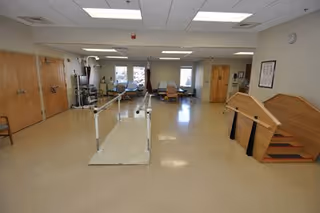 A spacious rehabilitation room with parallel bars for walking exercises, exercise equipment, chairs, and a wooden staircase with handrails for physical therapy. The room has a tiled floor, fluorescent ceiling lights, and windows letting in natural light.