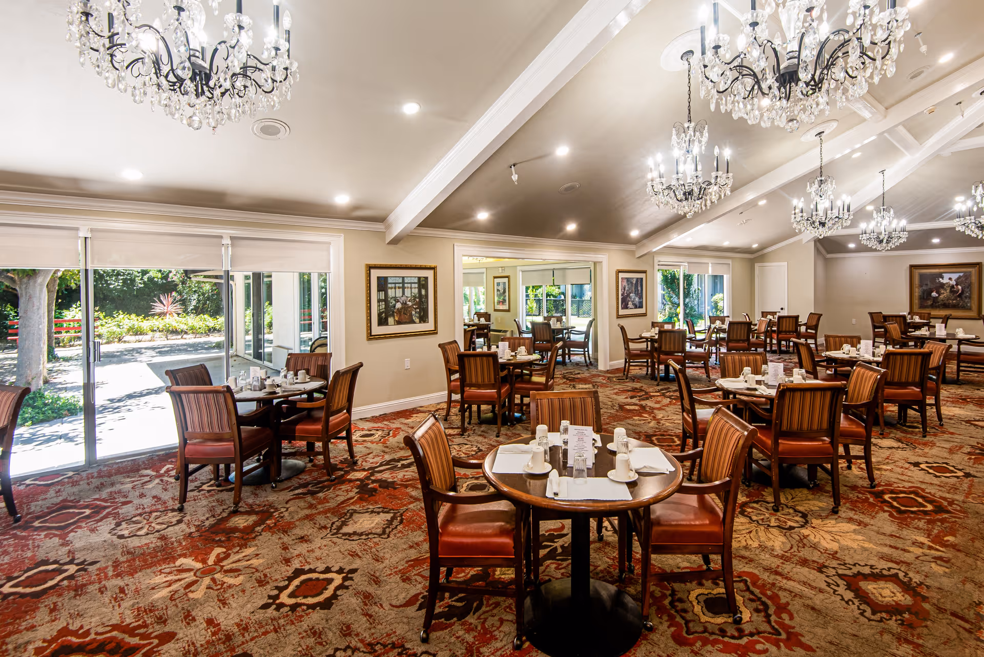 A spacious dining room with multiple round tables set with white cups, plates, and napkins. The room features large windows letting in natural light and offering views of a garden outside. The ceiling has several elegant chandeliers, and the floor is covered with a patterned carpet. Framed artwork decorates the walls.