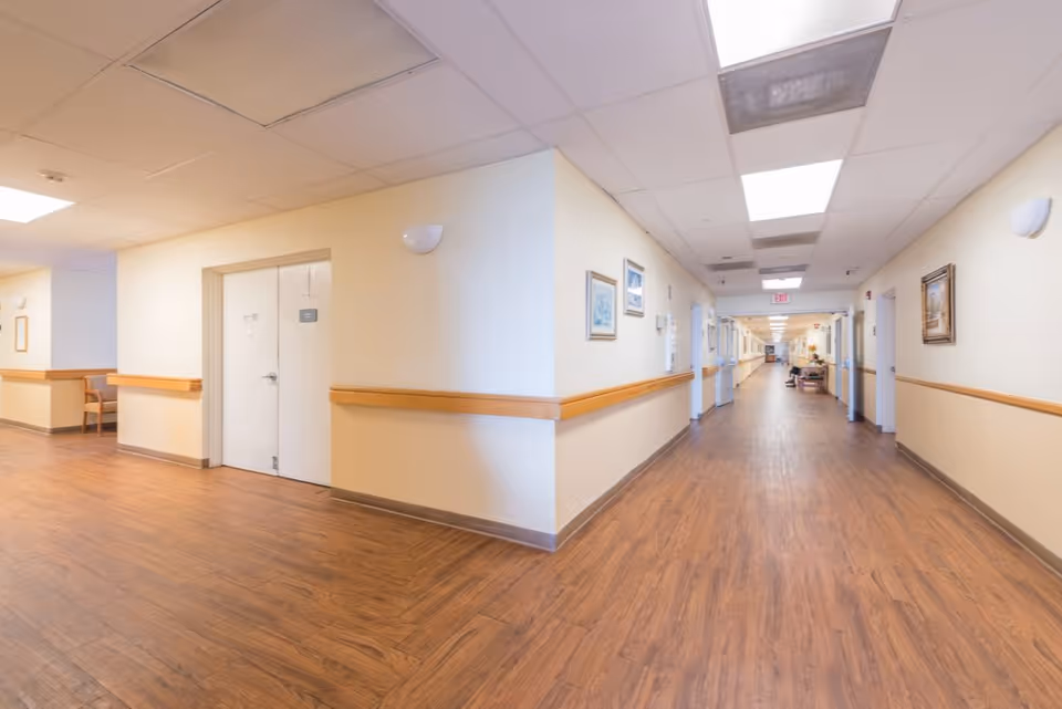 A long, well-lit hallway in a senior living facility with wooden flooring, cream-colored walls, handrails, framed pictures on the walls, and a few chairs placed along the corridor.