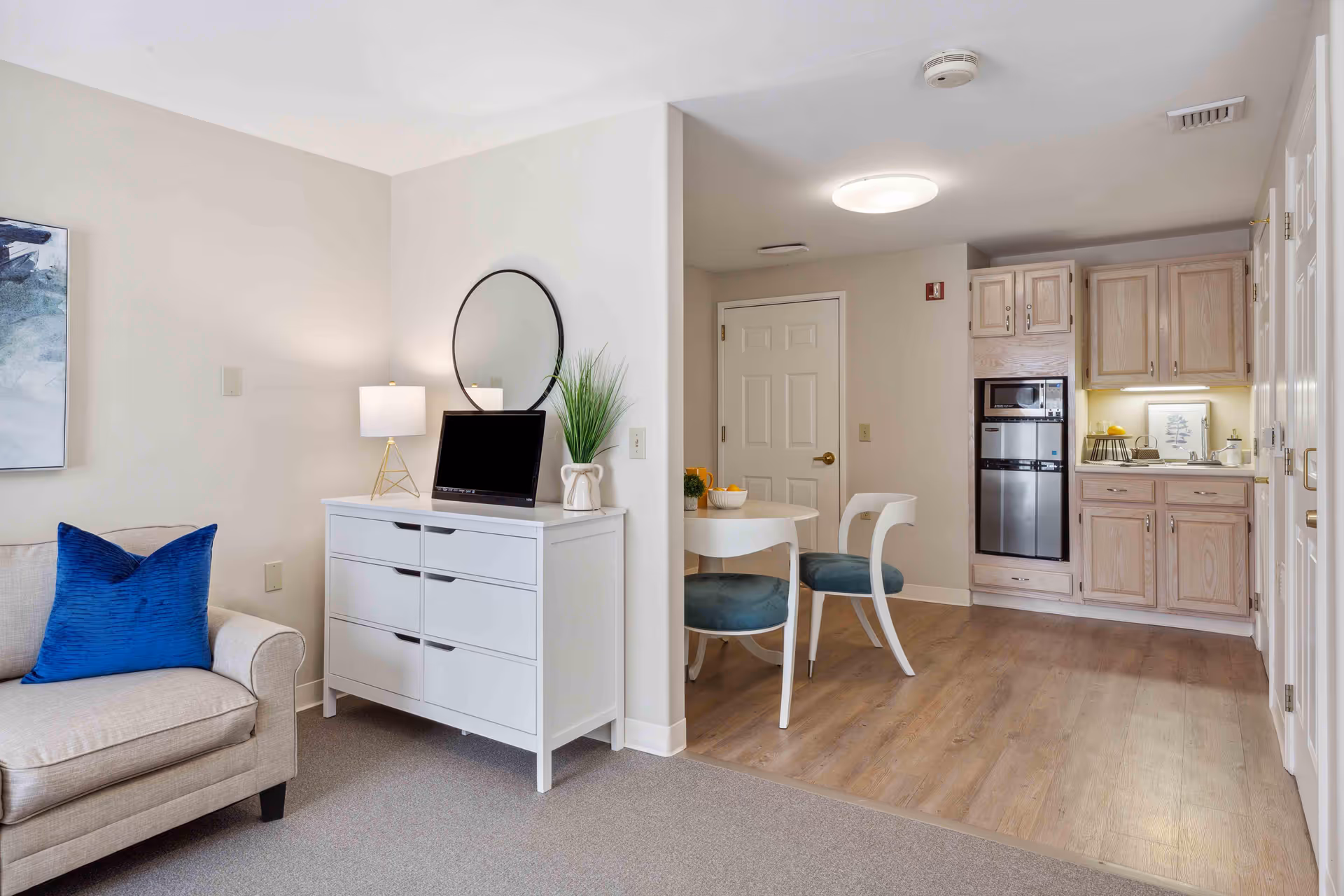 A cozy interior space featuring a beige armchair with a blue pillow, a white dresser with a round mirror, a small lamp, and a plant. Adjacent to the dresser is a dining area with a round table and two white chairs with blue cushions. The kitchen area has light wood cabinets, a built-in microwave, and an oven. The flooring transitions from carpet in the living area to wood in the dining and kitchen areas.