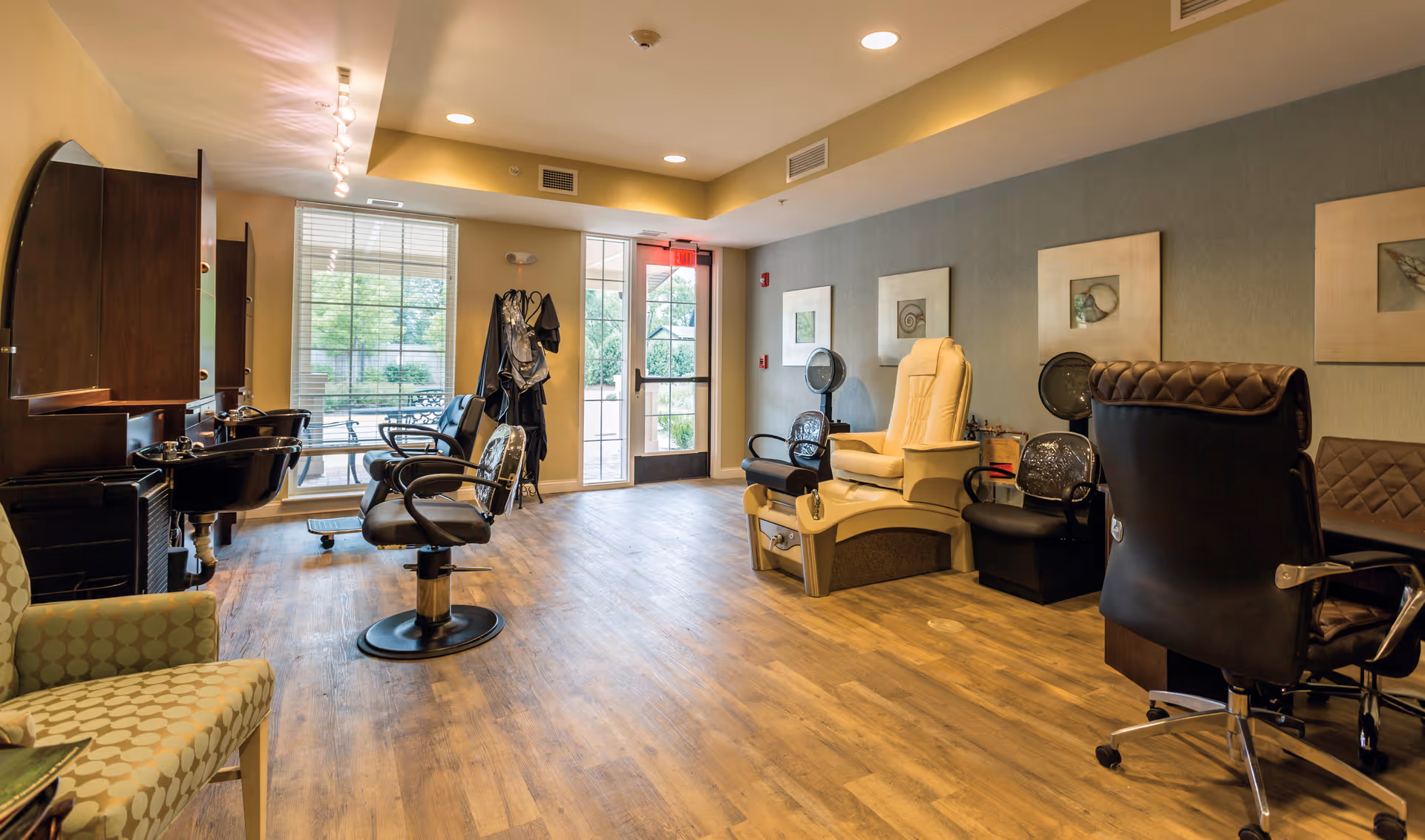 Interior view of a salon area in Cedar Lake Assisted Living and Memory Care featuring salon chairs, a pedicure chair, hair washing stations, and a seating area with large windows and a door leading outside.