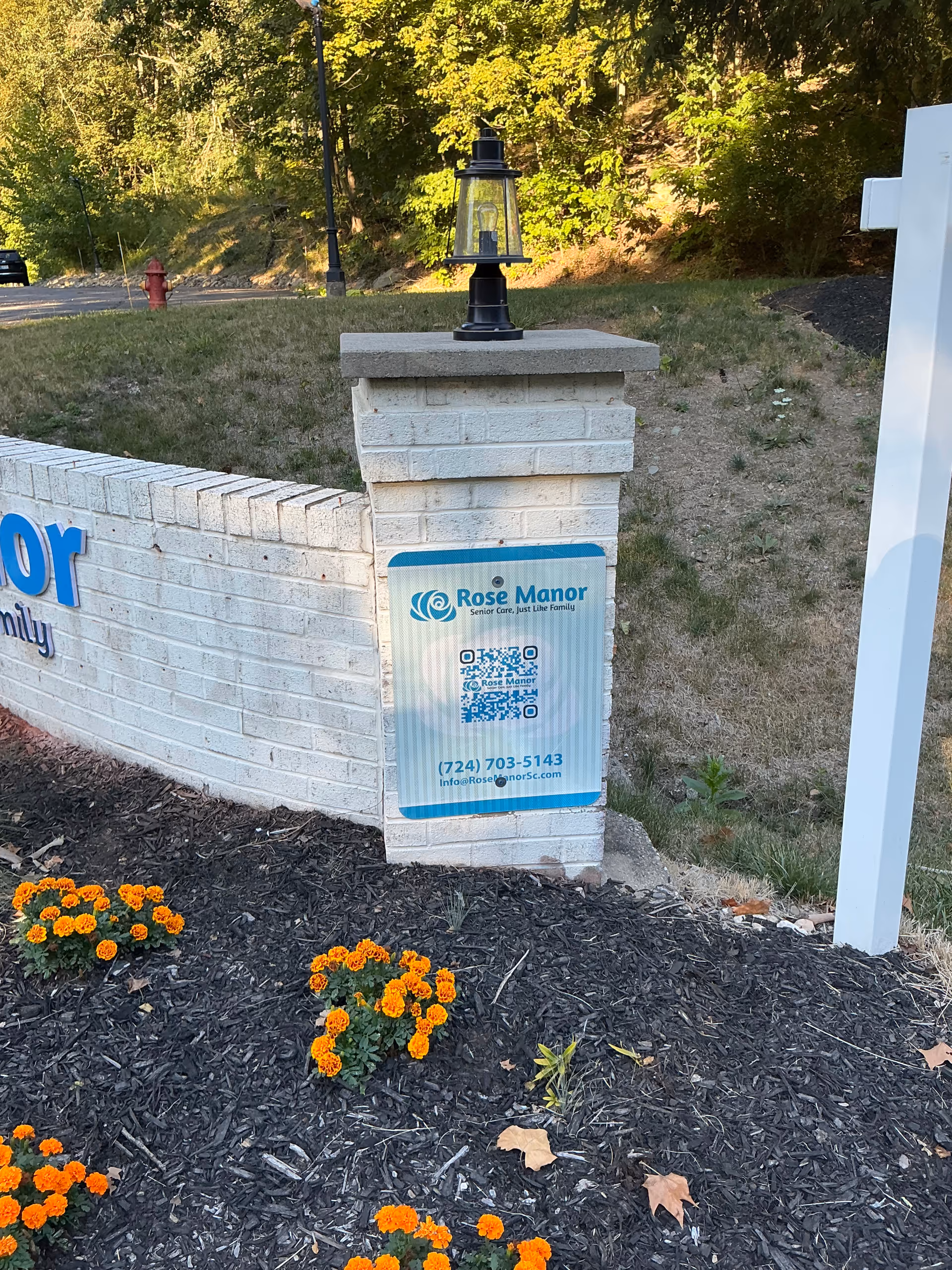 White brick entrance pillar topped with a lamp displaying a 'Rose Manor' sign beside a flower bed of orange marigolds and a grassy slope.
