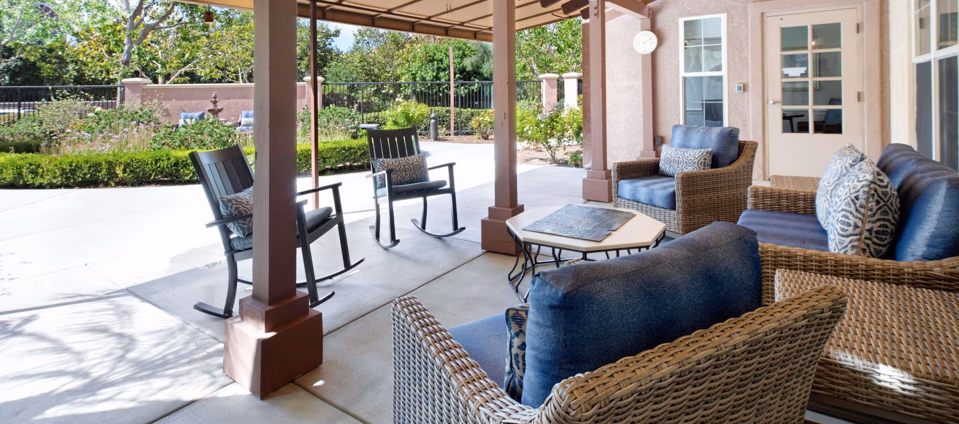 Covered outdoor patio area with wicker furniture including cushioned chairs and a sofa with blue cushions and patterned pillows, two wooden rocking chairs with cushions, a small hexagonal table with a puzzle on top, and a view of a garden with greenery and a fountain in the background.
