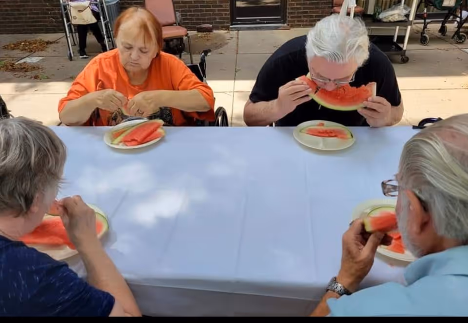 Four elderly individuals sitting around a table outdoors, eating slices of watermelon. The table is covered with a white tablecloth, and the setting appears to be a patio area with brick walls and chairs in the background.