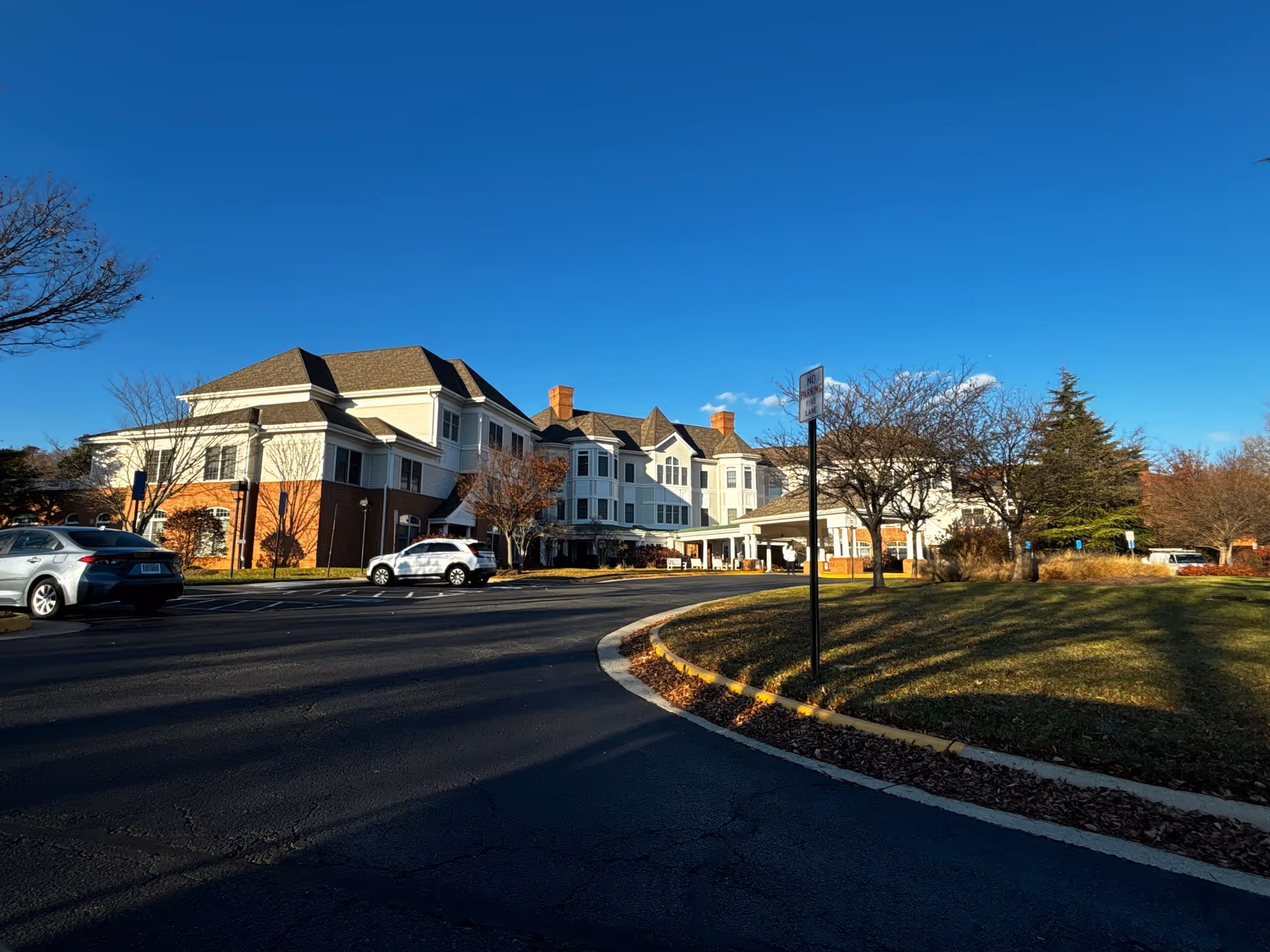 Exterior view of a large senior living facility building with white and brick facade under a clear blue sky. There are a few cars parked along the driveway and some leafless trees around the building.