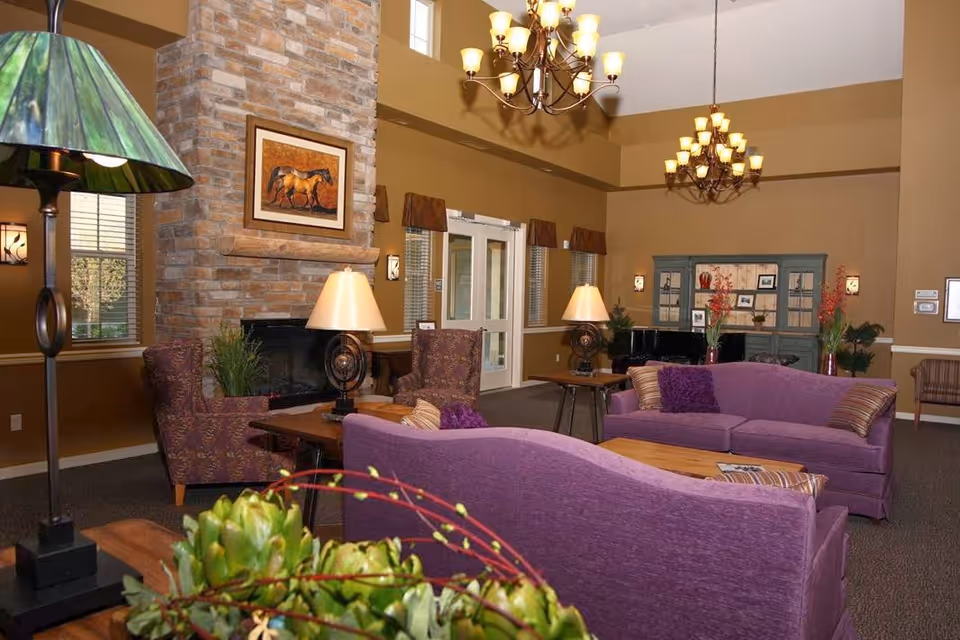 Comfortable communal living room with purple sofas, armchairs, a stone fireplace, chandeliers, and decorative plants.