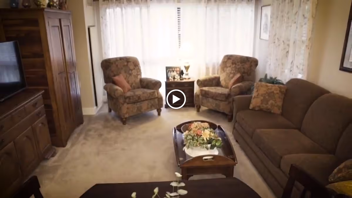 A cozy living room with two floral-patterned armchairs and a brown sofa with matching cushions. A wooden coffee table with a floral centerpiece is in the center, and a wooden cabinet with a TV is on the left side. Sheer curtains cover the windows, allowing soft natural light to fill the room.