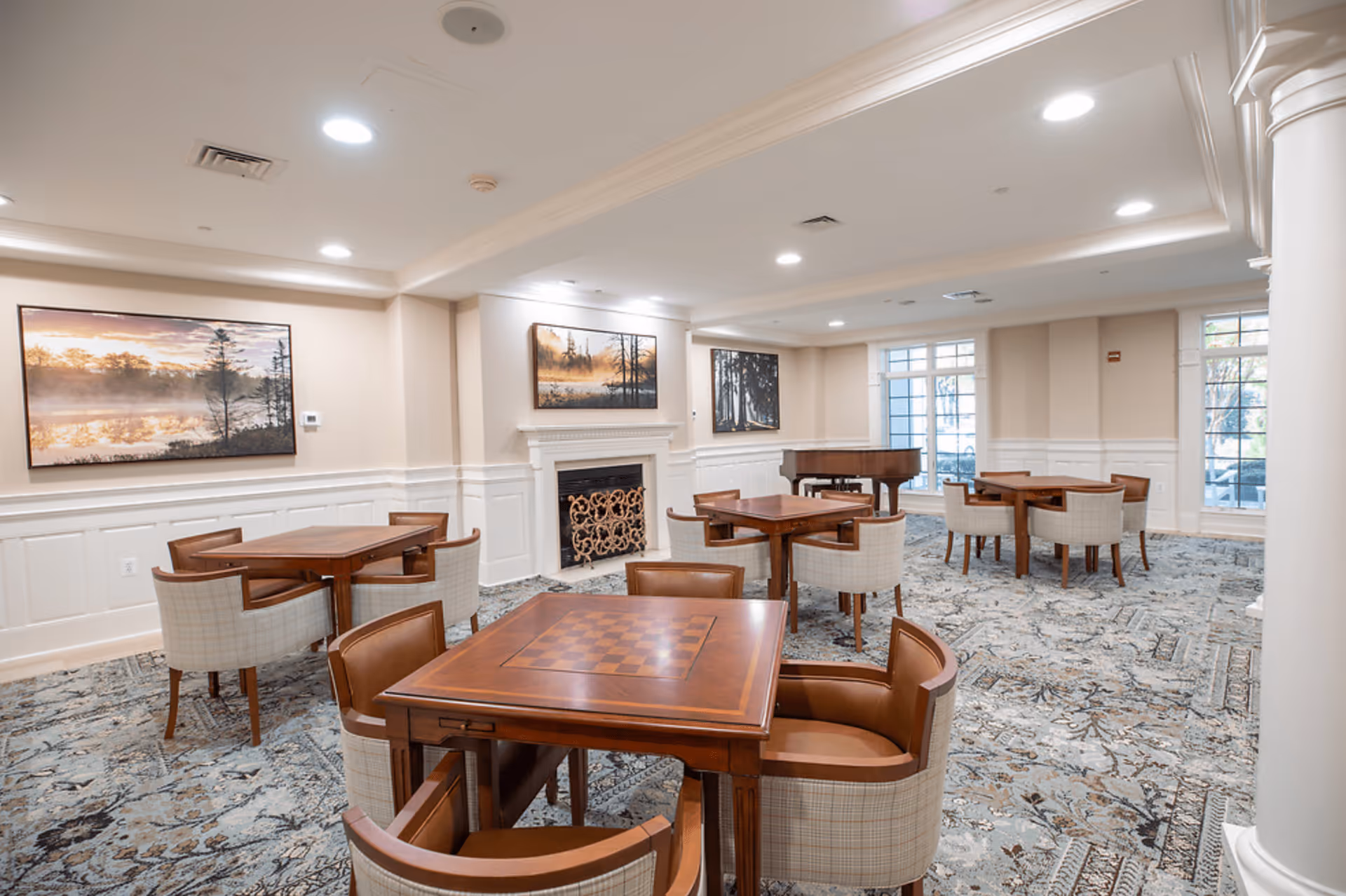 A well-lit common room with several wooden tables and cushioned chairs arranged around them. The room features a fireplace with decorative screen, large windows letting in natural light, and framed landscape artwork on the walls. A grand piano is positioned near the windows.