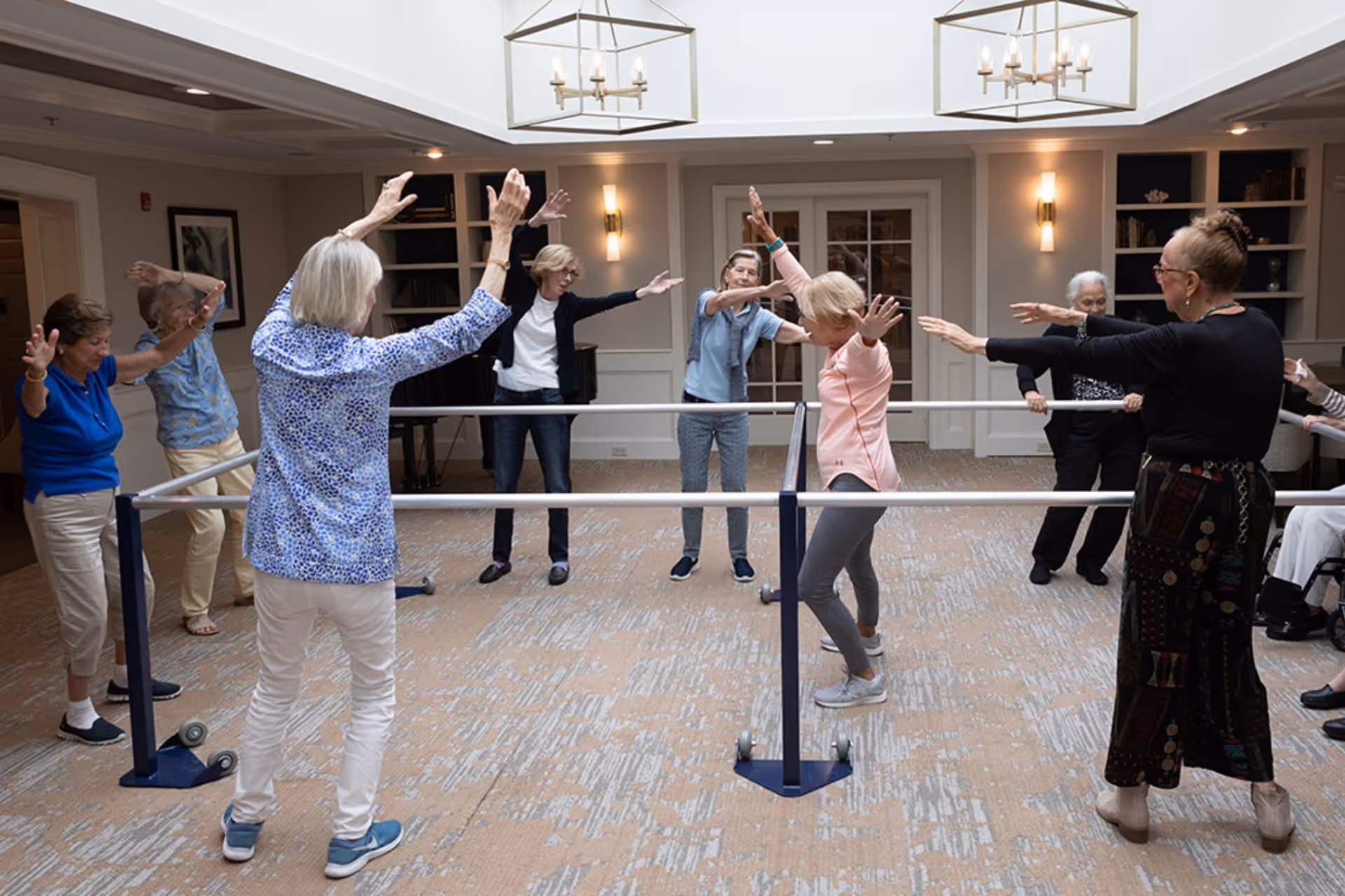 A group of elderly women participating in a standing exercise class using parallel bars in a spacious, well-lit room with carpeted floors and modern light fixtures.