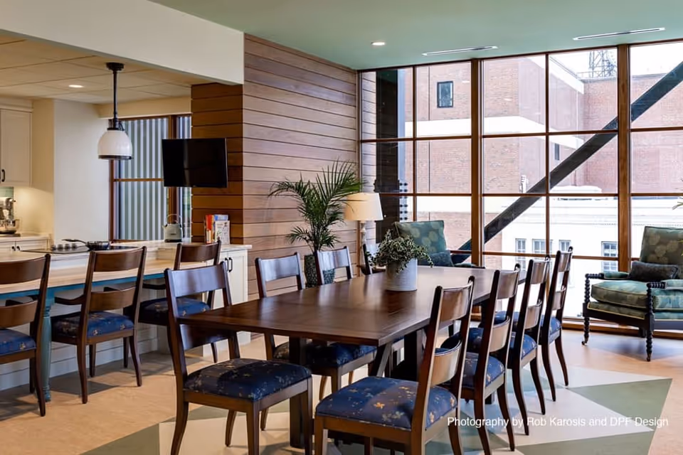 A spacious dining area with a large wooden table surrounded by wooden chairs with blue cushioned seats. To the left, there is a kitchen counter with additional chairs and a hanging light fixture above. Large floor-to-ceiling windows reveal an exterior view of brick buildings and a metal staircase. The room is decorated with a potted plant on the table and another plant near the window, along with two upholstered armchairs with patterned cushions near the windows.