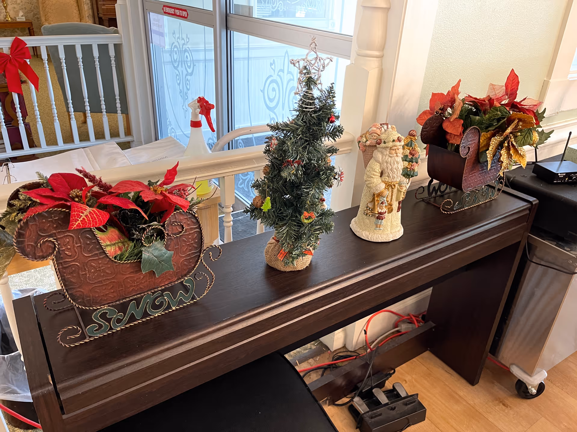A dark wooden table decorated with Christmas-themed items including two red sleighs filled with poinsettias, a small decorated Christmas tree, and a Santa Claus figurine. In the background, there is a white railing with a red bow and a glass door with decorative frosted patterns.