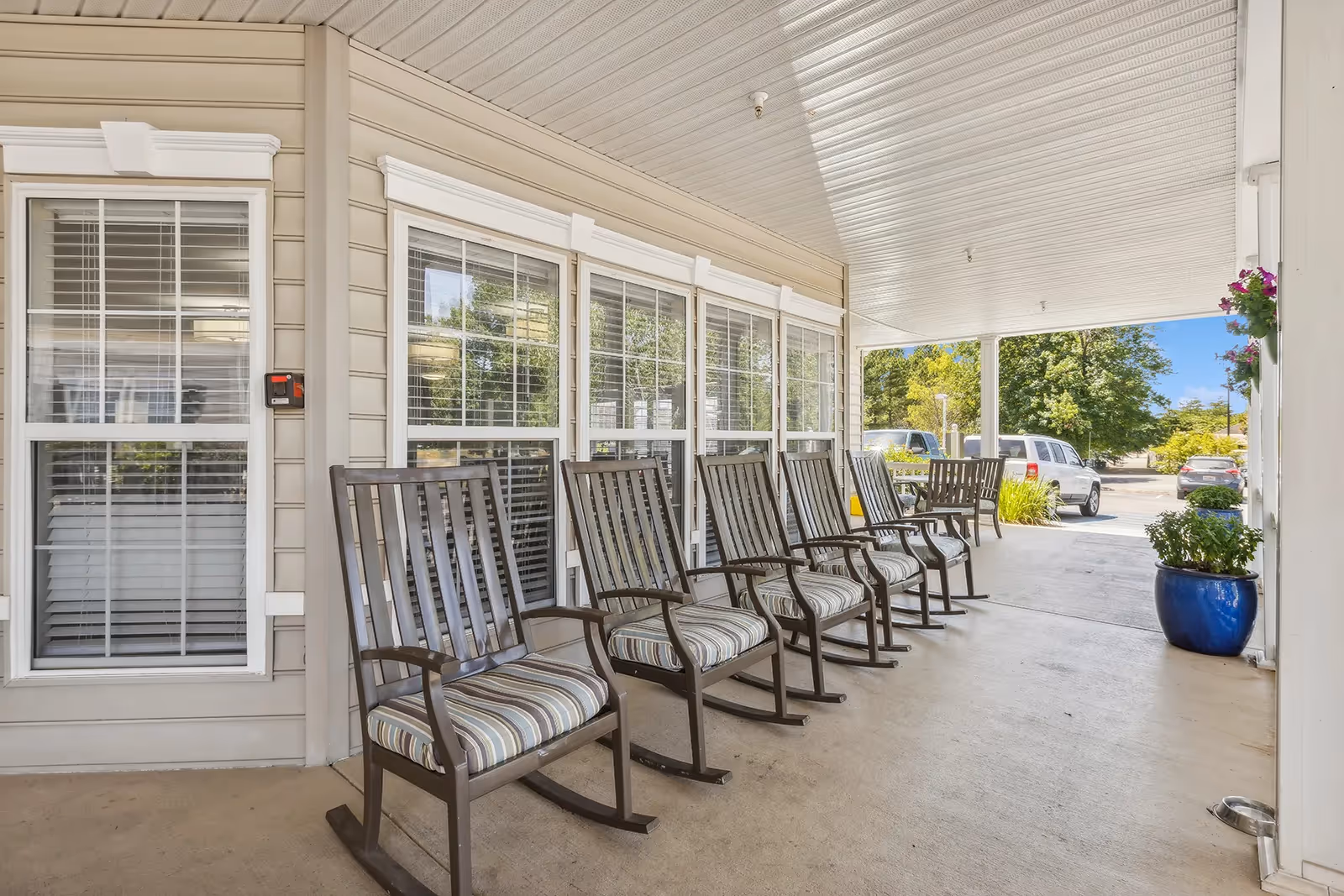 Covered front porch with a row of rocking chairs lined up in front of windows and potted plants overlooking the driveway.