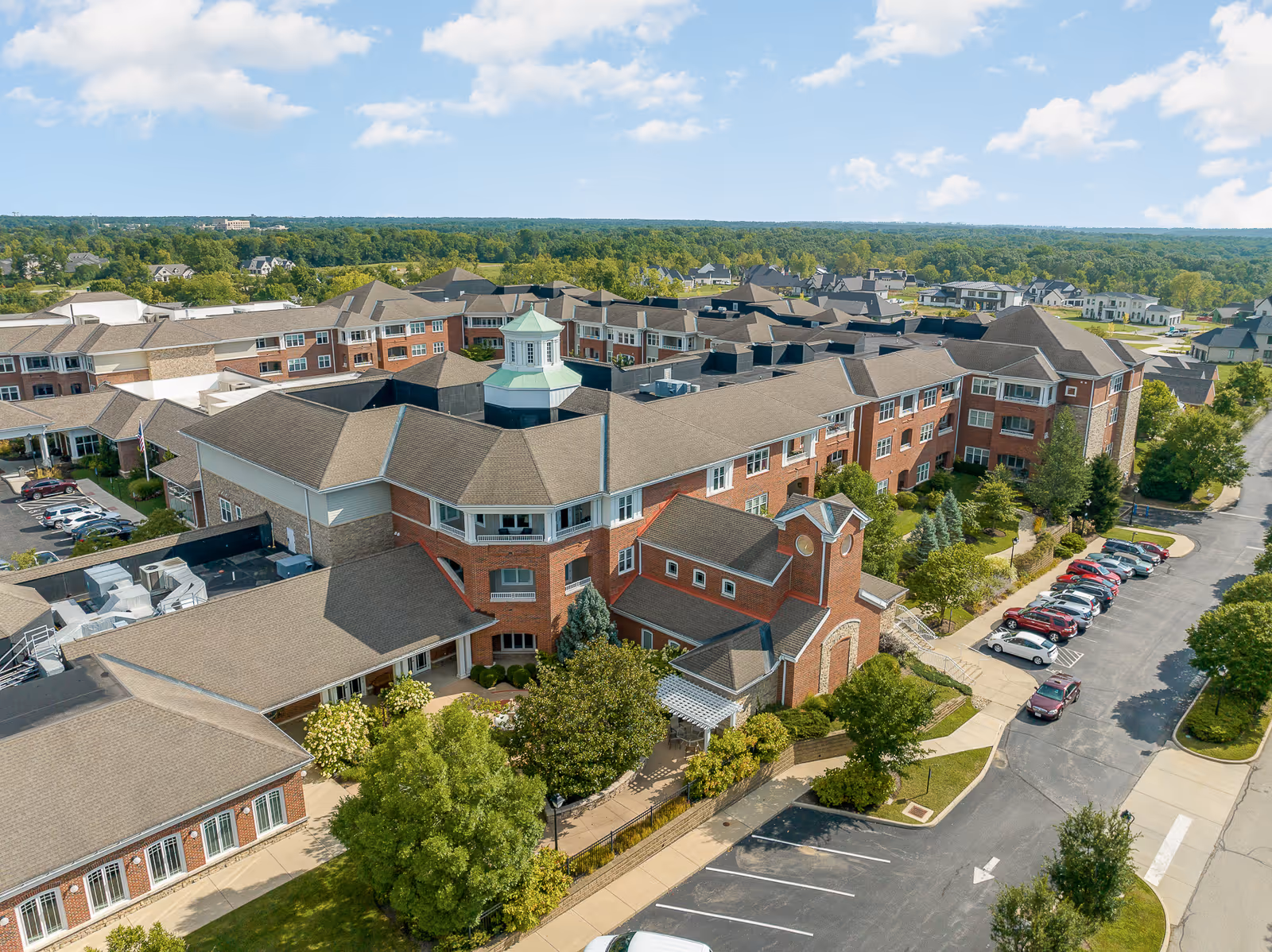 Aerial view of a large red-brick senior living complex with multiple rooflines, trees, and a parking lot.