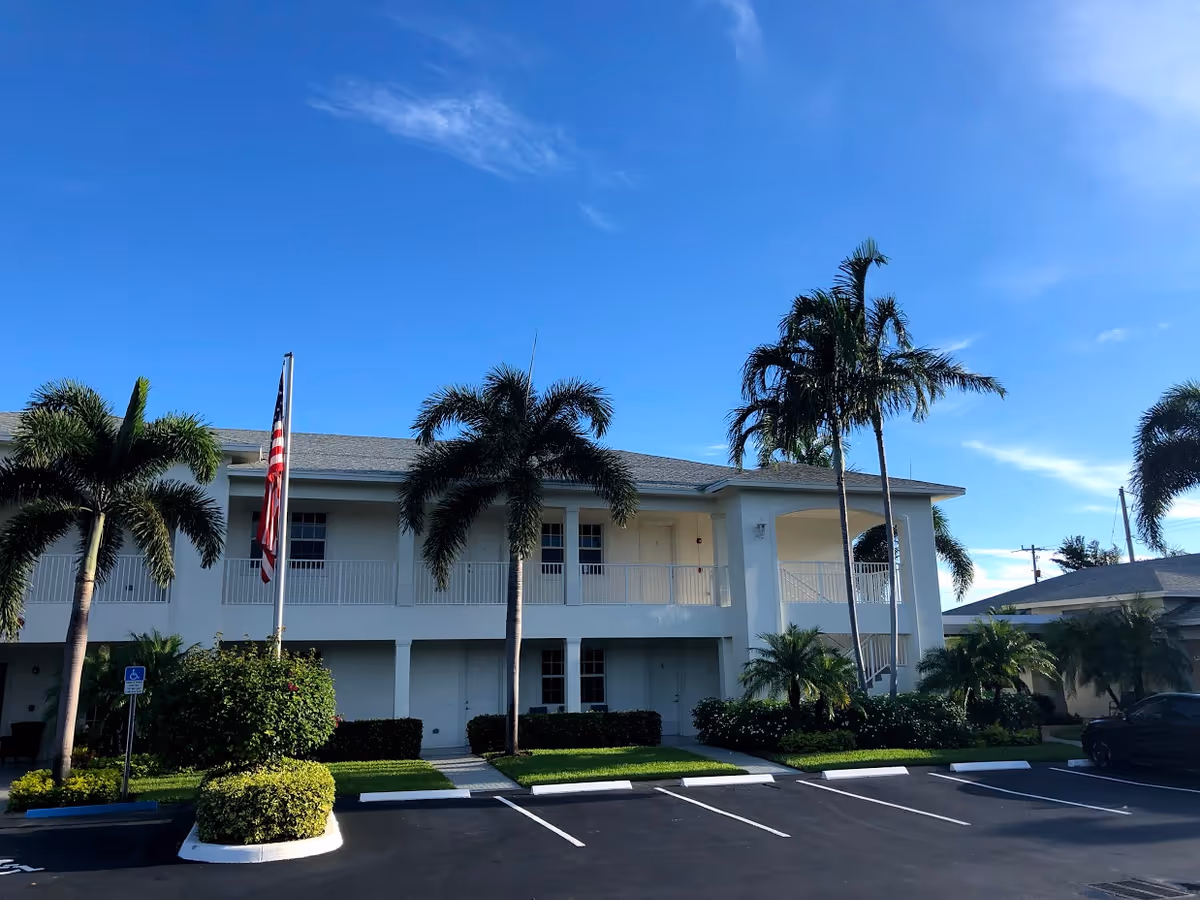 Exterior view of a two-story white building with a balcony and several palm trees in front. There is an American flag on a flagpole near the center, and a parking lot with marked spaces in the foreground under a clear blue sky.