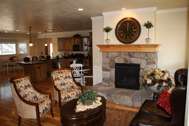 Cozy living room area with a stone fireplace topped by a large round clock and two small potted plants. The room features two floral-patterned armchairs, a dark leather sofa with red pillows, a wooden coffee table with a plant centerpiece, and an open view into a kitchen and dining area with wooden cabinetry and hardwood floors.