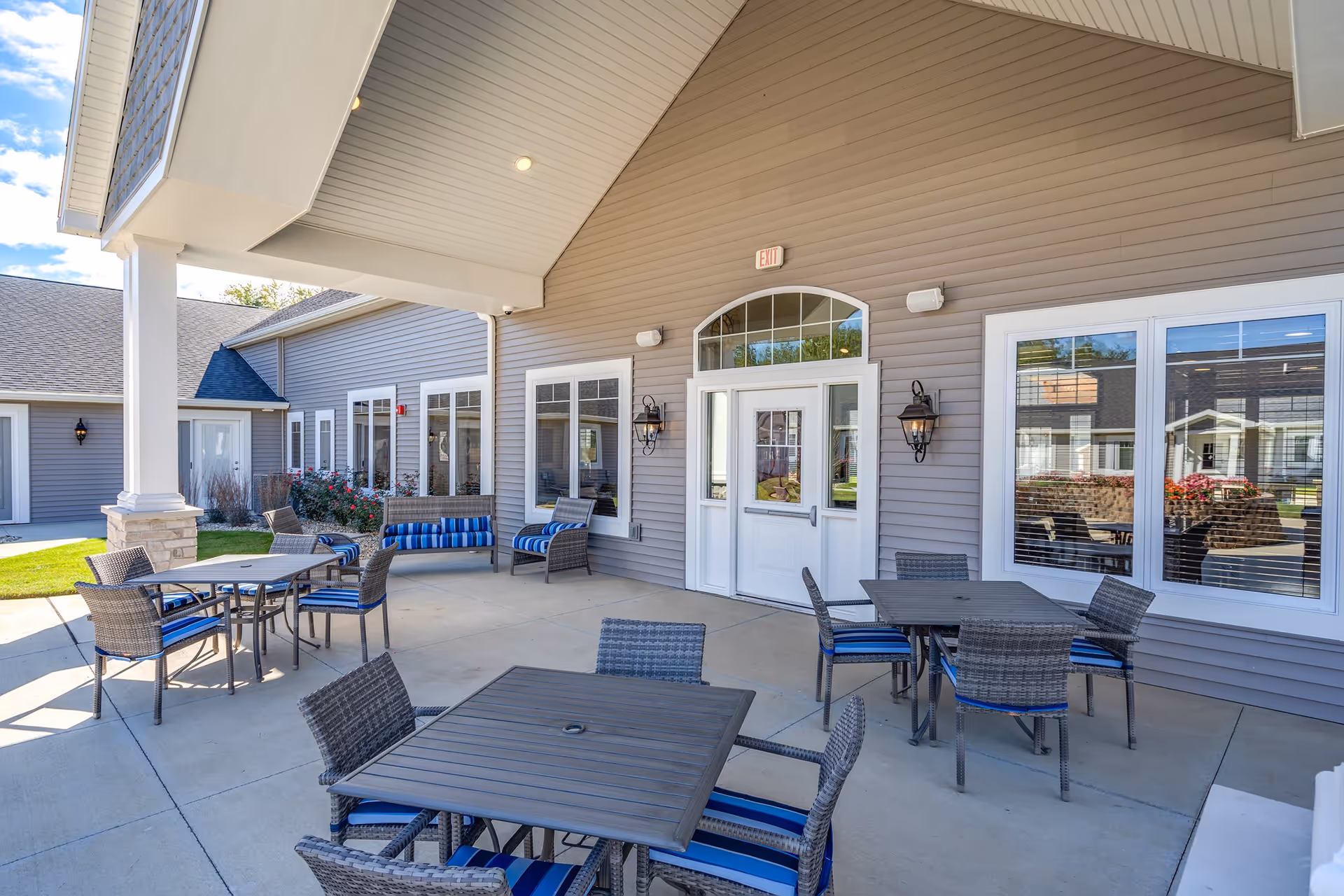 Covered outdoor patio with multiple tables, wicker chairs and benches in front of a senior living facility entrance.