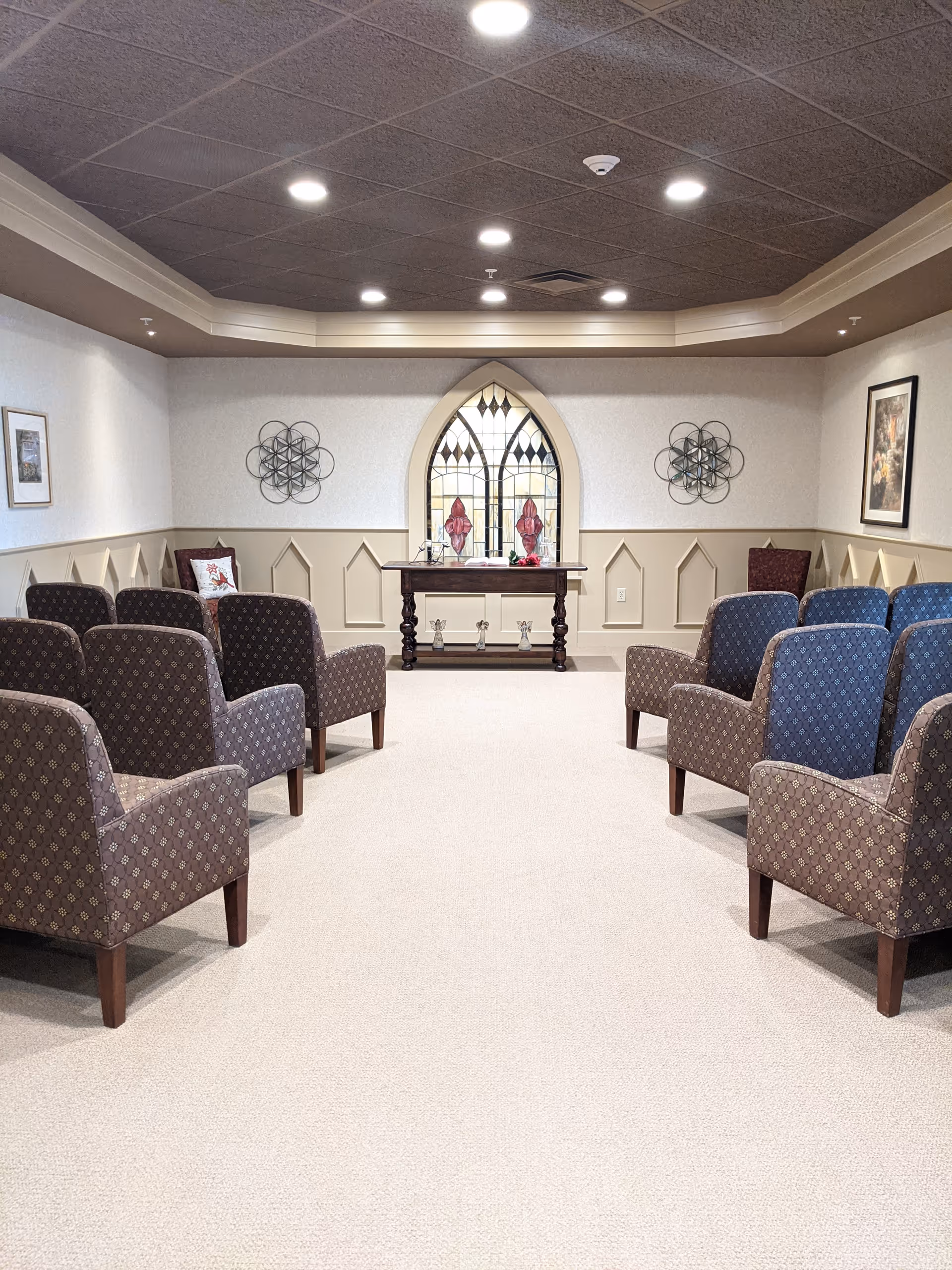 A small chapel or meditation room with rows of patterned armchairs facing a wooden table in front of a stained glass window. The room has beige walls with decorative paneling, framed artwork, and ceiling lights.