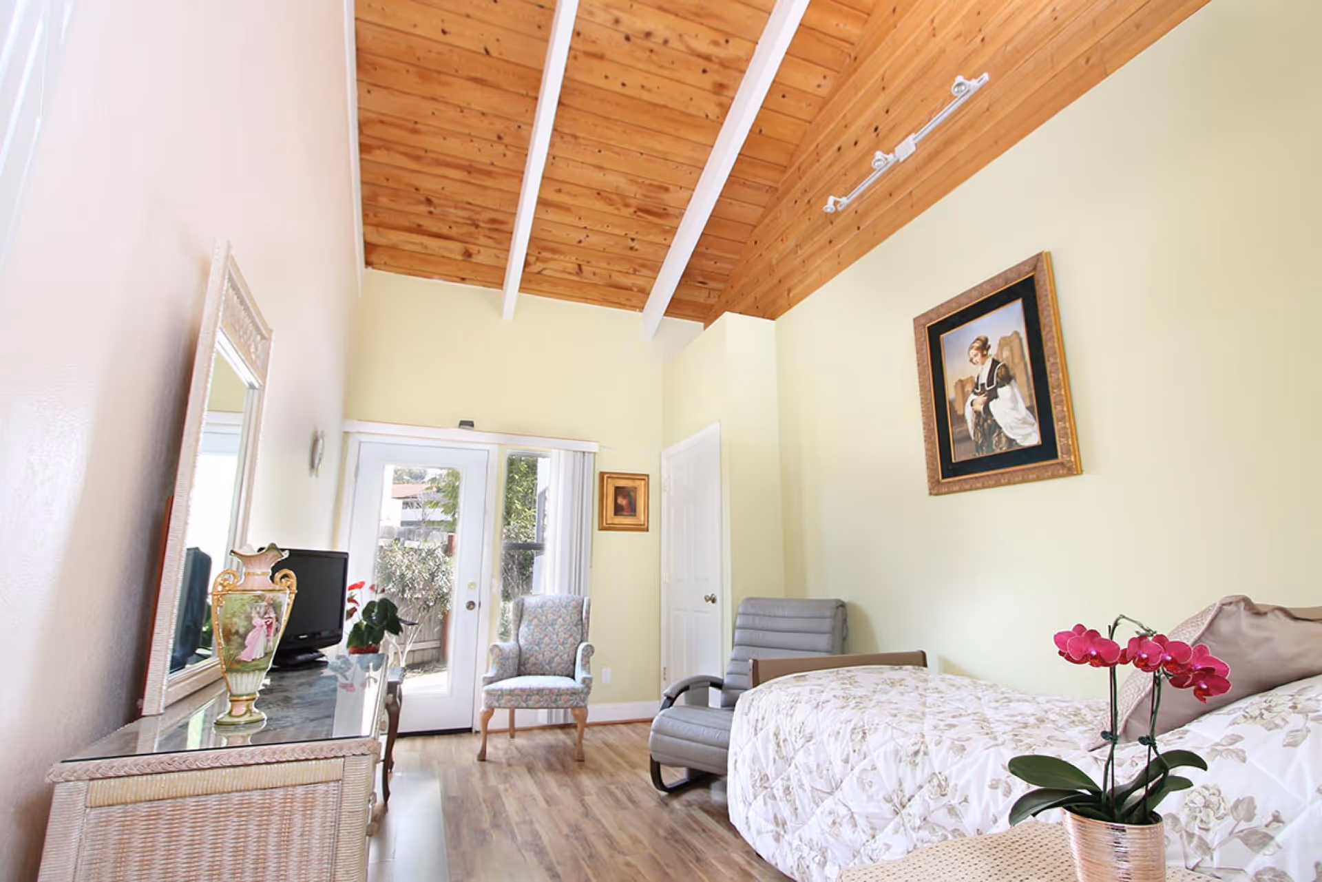 A cozy bedroom with a wooden vaulted ceiling and light yellow walls. The room features a bed with a floral quilt, a small table with a pink orchid plant, a gray armchair, and a floral upholstered chair near glass doors leading outside. A dresser with a decorative vase and a mirror is on the left side, and a framed painting hangs on the wall above the bed.