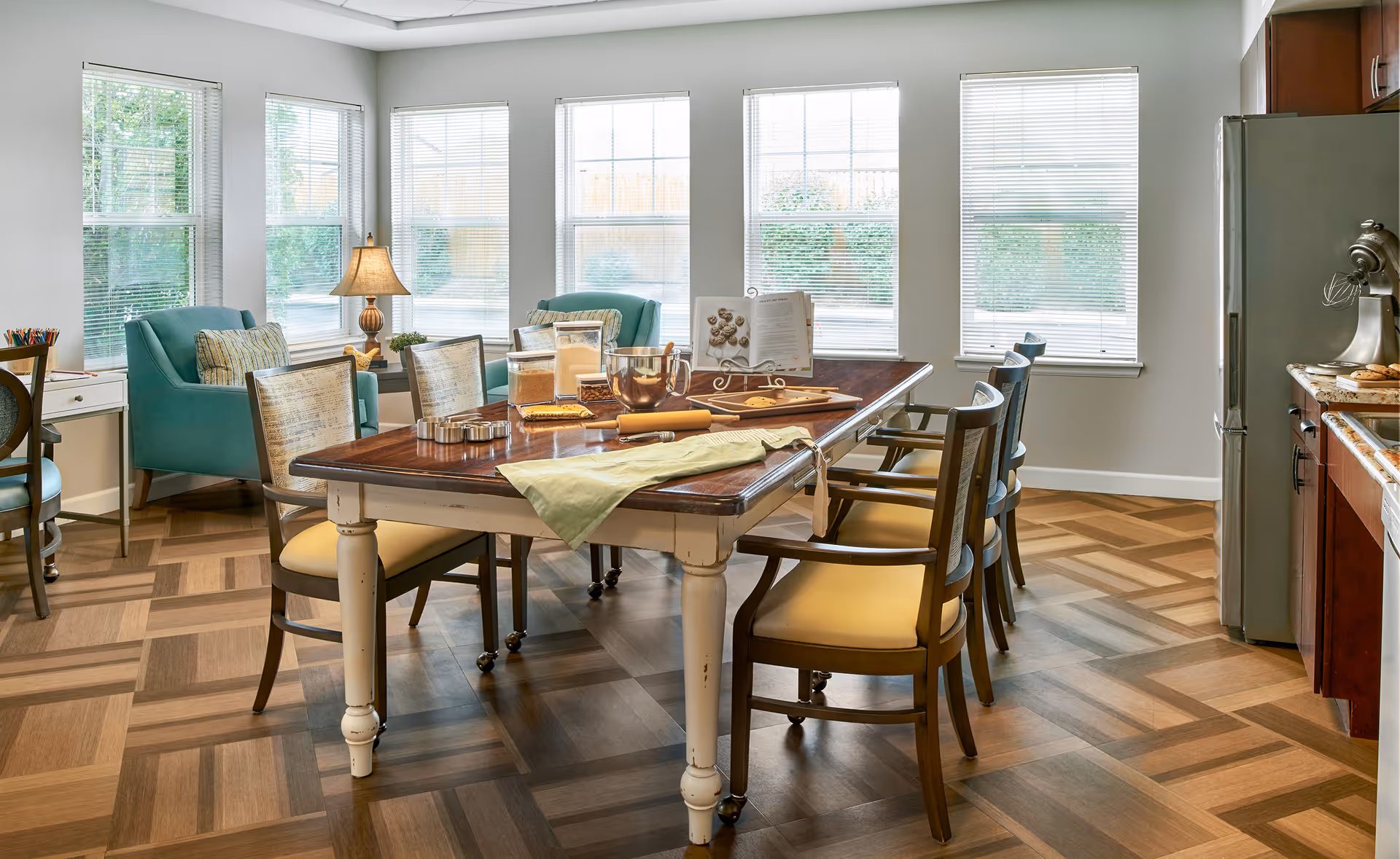 A bright dining area with a wooden table set for baking, including a rolling pin, cookie cutters, and ingredients. The room has large windows with blinds, two teal armchairs, a lamp, and a kitchen area with a refrigerator and mixer visible.