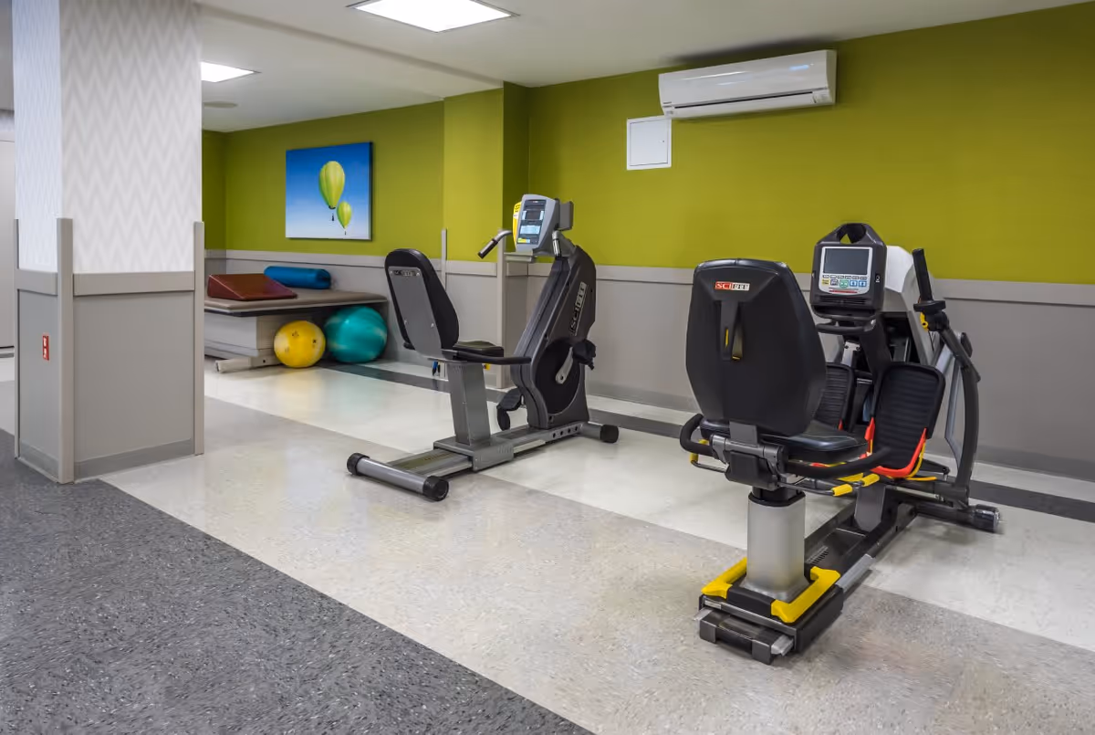 A small exercise area in a rehabilitation and healthcare center featuring two recumbent exercise bikes, a bench with exercise balls and a foam roller, and a green accent wall with a painting of two hot air balloons.