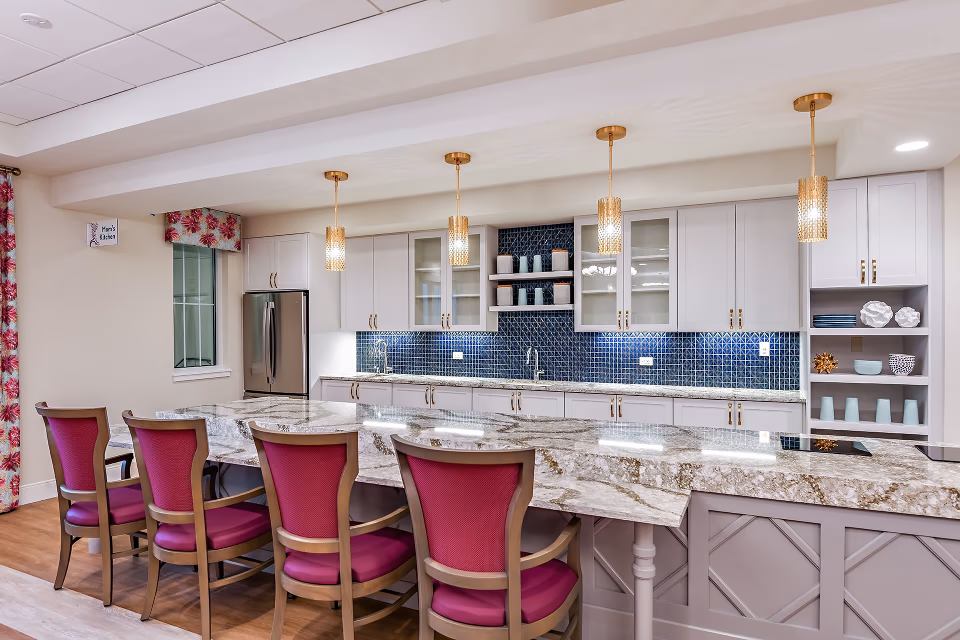 Bright community kitchen with a large marble island, pendant lights, and a row of magenta-upholstered bar chairs.