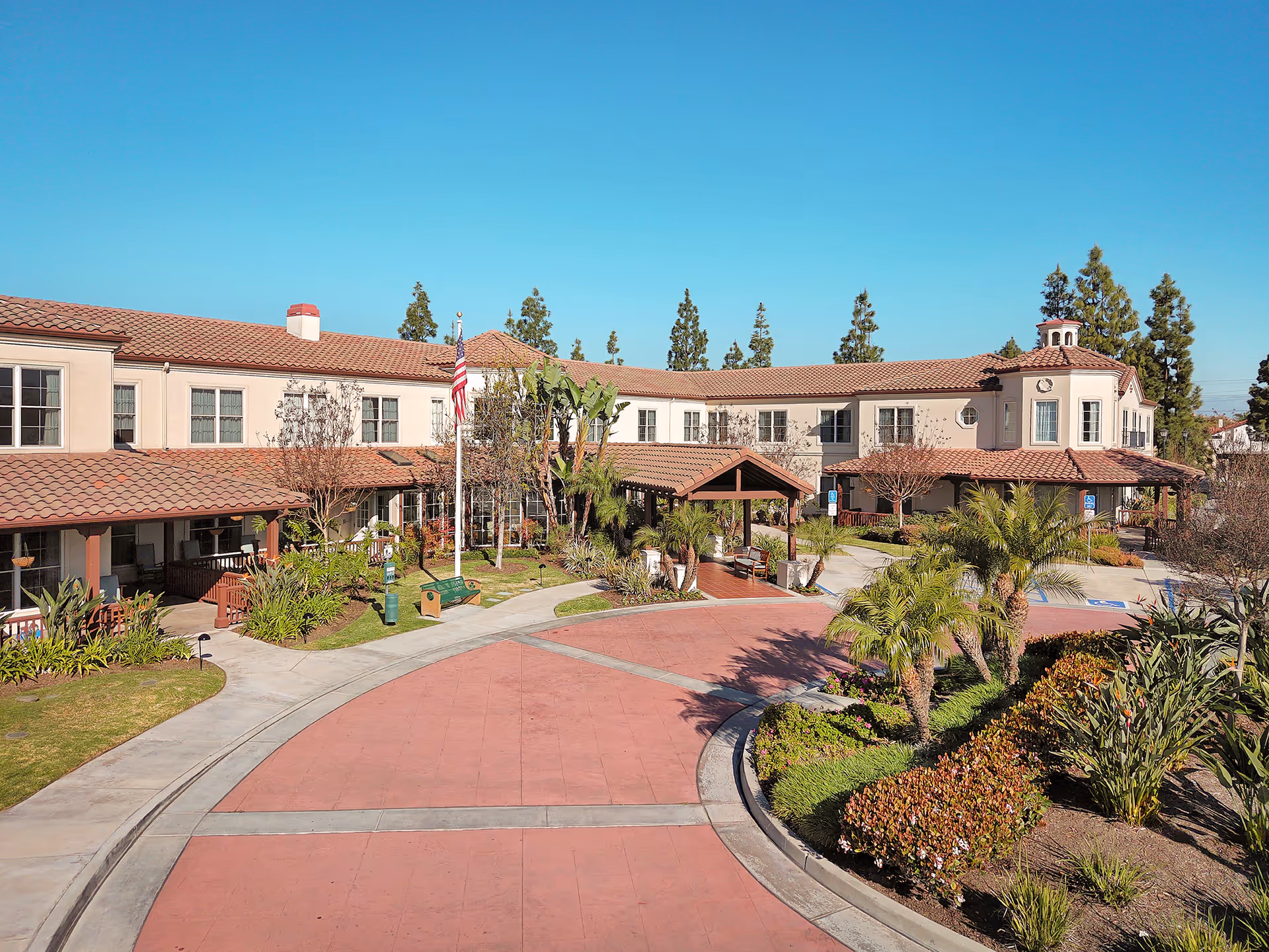 Exterior view of a senior living facility named Ivy Park at La Palma, featuring a two-story building with a tiled roof, an American flag on a flagpole, landscaped gardens with palm trees and shrubs, and a curved driveway with parking spaces.