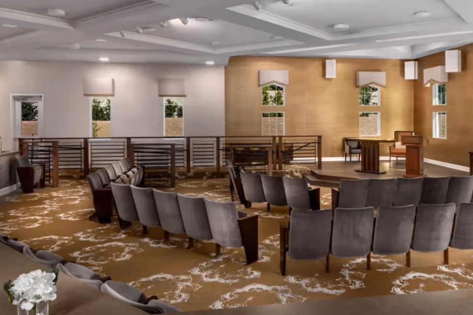 A spacious room with rows of gray upholstered chairs arranged in a semi-circle facing a small wooden podium and table on a raised platform. The room has beige walls with several windows covered by valances, a patterned carpet, and a white ceiling with recessed lighting.