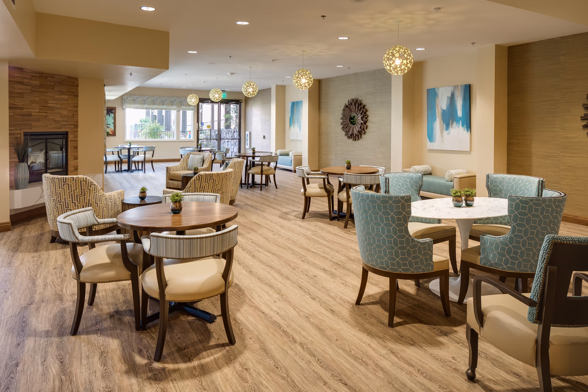 A spacious, well-lit common area with multiple round tables and chairs arranged for seating. The room features wooden flooring, modern pendant lights hanging from the ceiling, a fireplace on the left side, and decorative wall art. The area appears comfortable and inviting, suitable for socializing or dining.